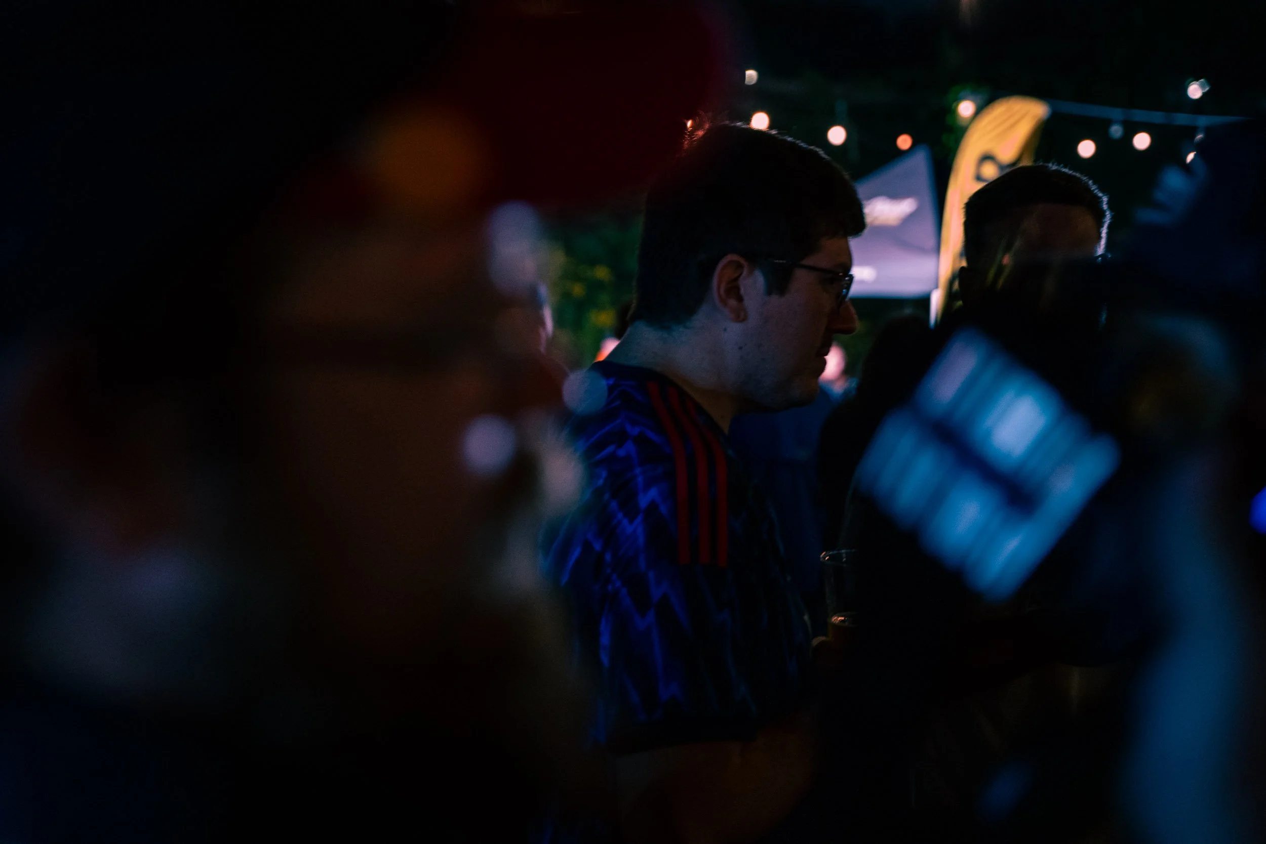 People at a nighttime outdoor event, some wearing glasses, with colorful lights and a flag in the background.