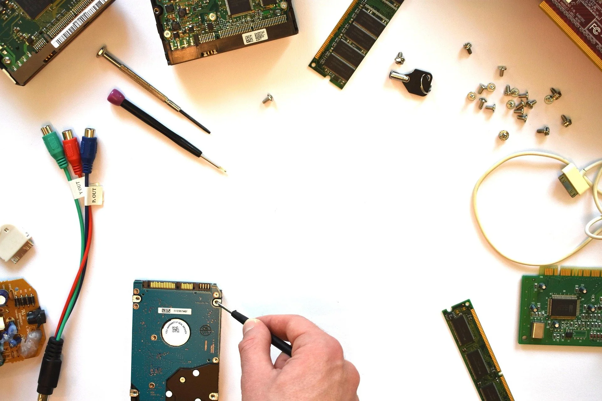 Person using a screwdriver to work on a blue hard drive surrounded by various computer components, circuit boards, RAM sticks, cables, and screws on a white surface.