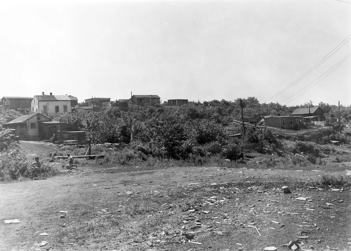Black and white photo of an impoverished neighborhood with some children playing outside, scattered small houses on a hill, and power lines overhead.