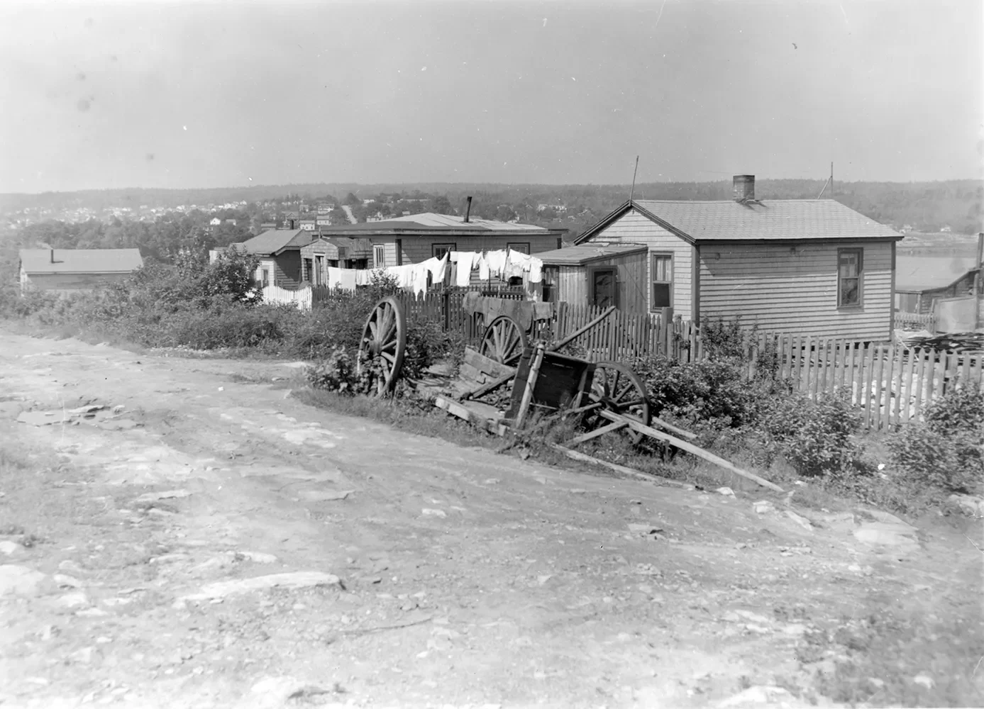 Black and white photograph of fenced houses on a dirt road and a broken wheelbarrow.