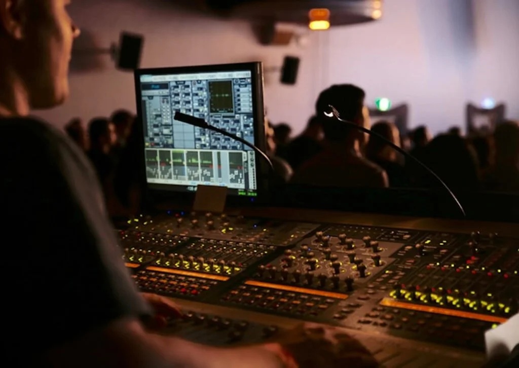 A sound engineer working on a digital audio mixing console with a computer screen displaying audio levels, in a crowded concert hall.