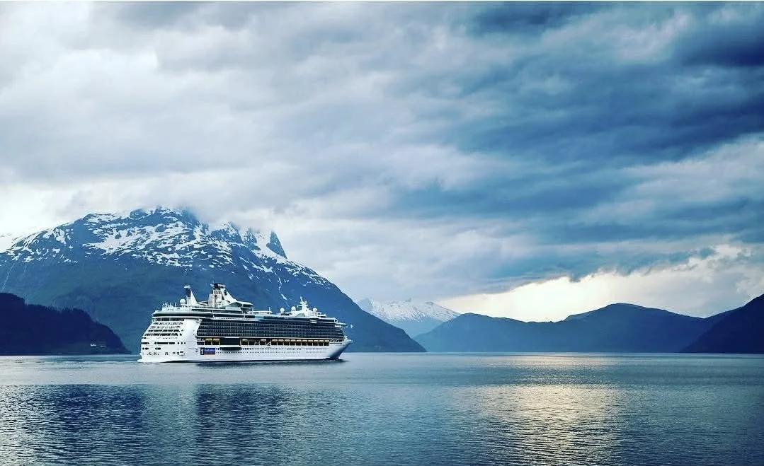A large cruise ship sailing through a fjord surrounded by snow-capped mountains and dramatic cloudy sky.