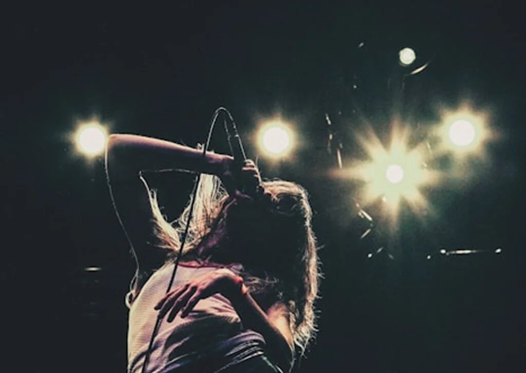 A woman singing into a microphone on stage with bright lights shining behind her.