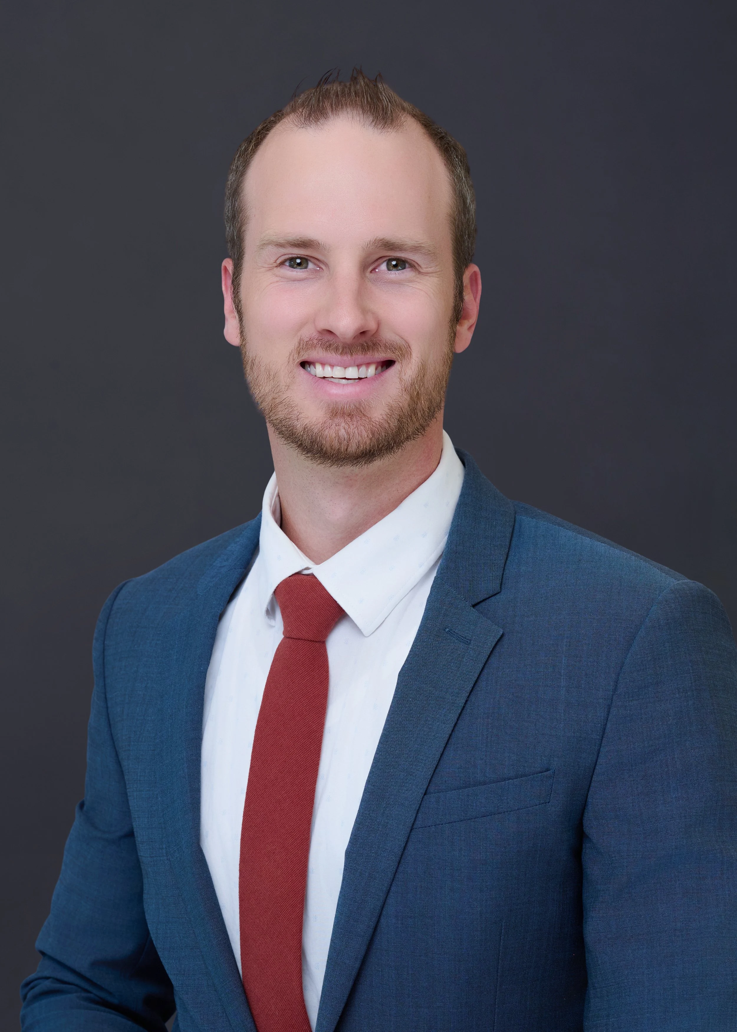 A young man with light skin, light brown hair, and a beard, smiling, wearing a dark blue suit, white shirt, and red tie, against a gray background.