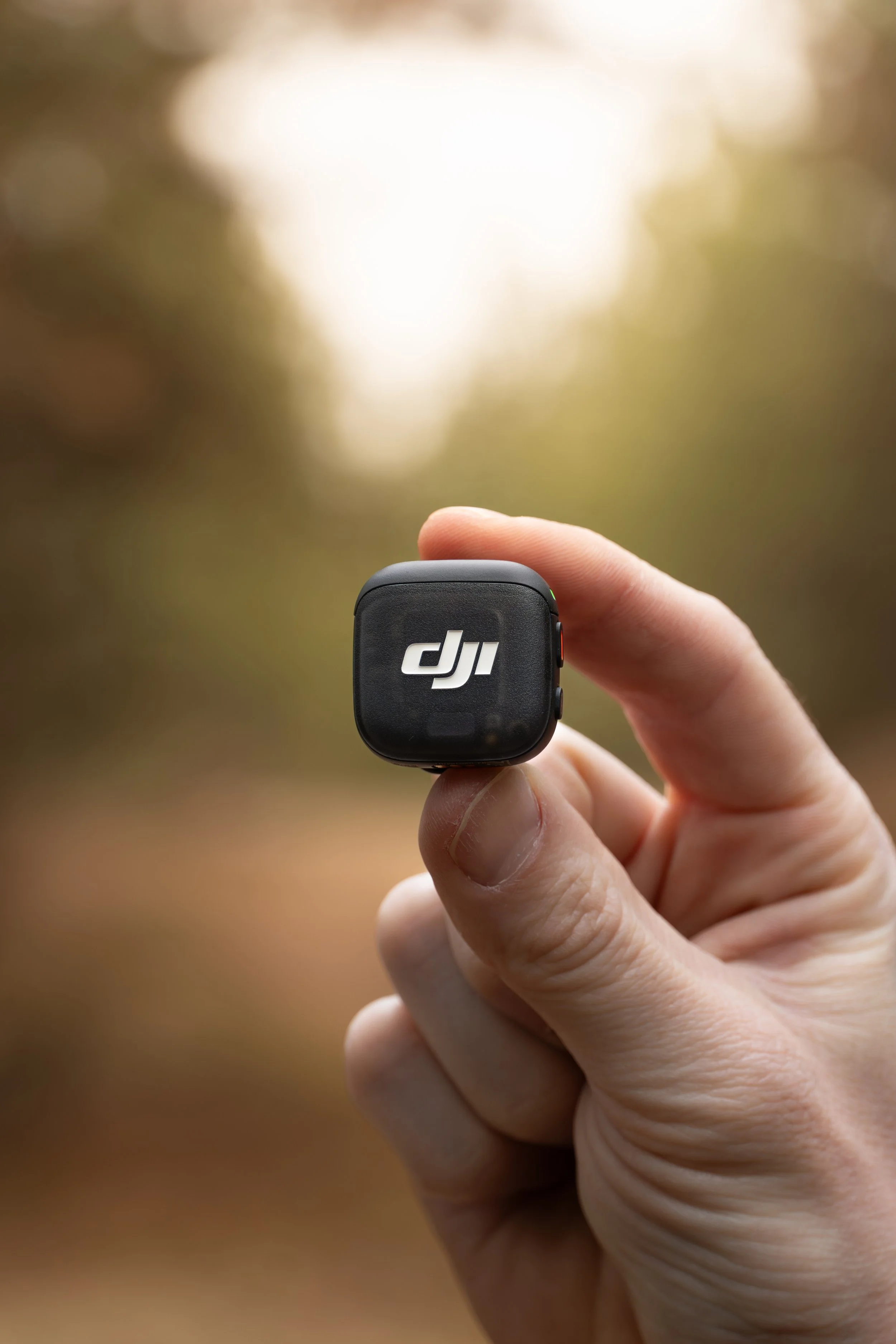 A hand holding a small black DJI device outdoors near trees with blurred sunlight in the background.