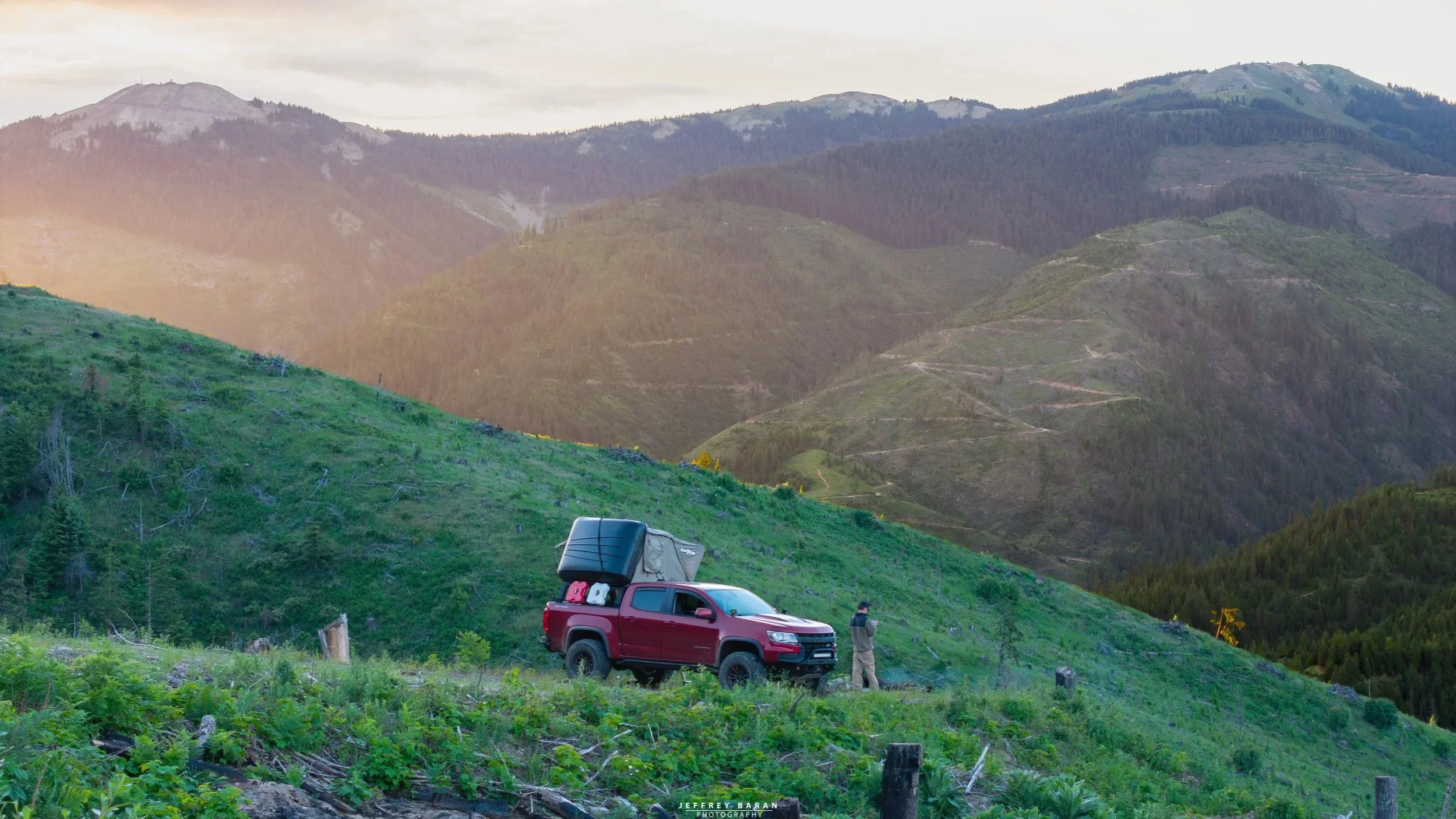 A mountainous landscape with green hills, trees, and valleys under a cloudy sky, featuring a red pickup truck with gear on the roof and two people standing nearby on a grassy slope.