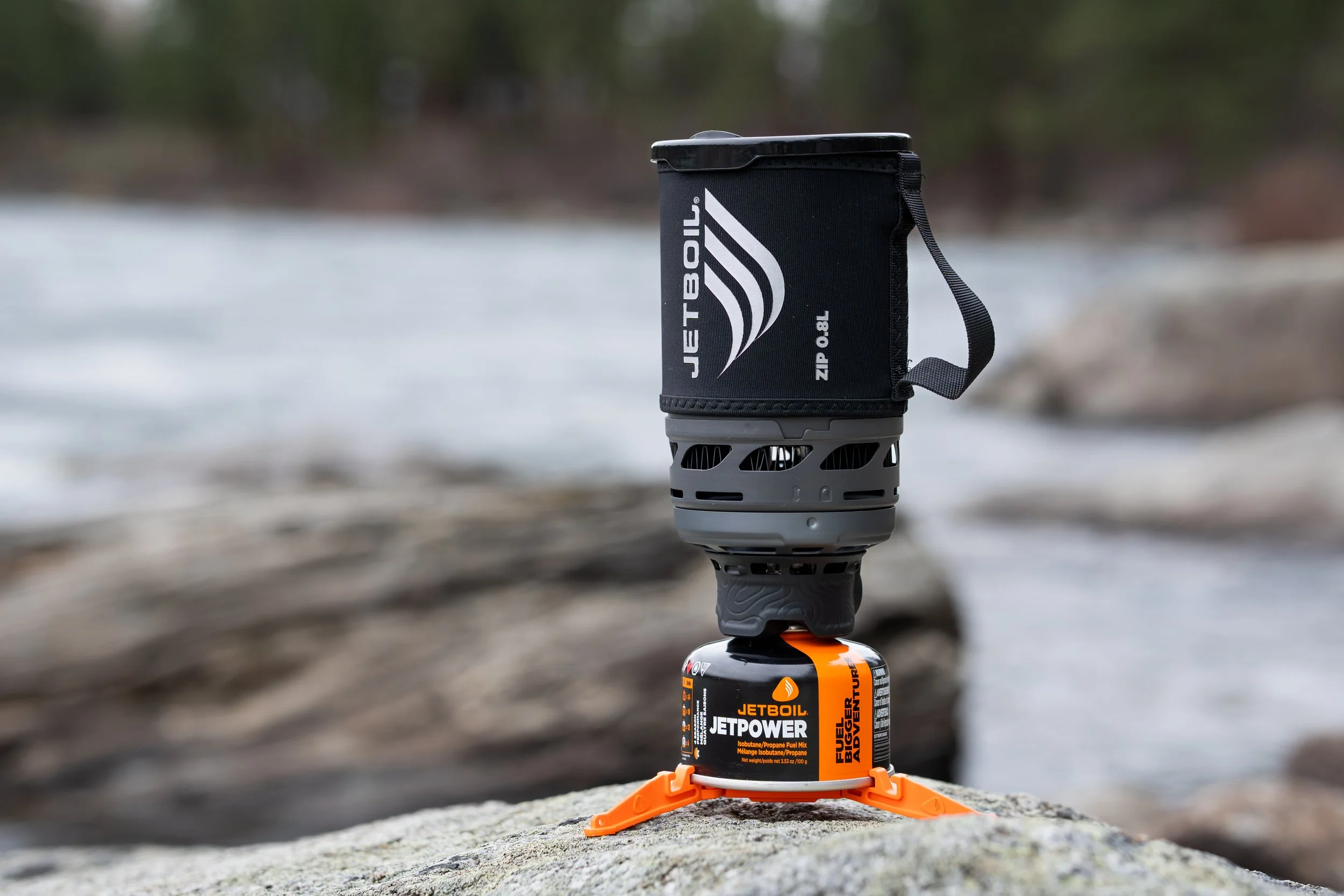 A Jetboil Jetpower fuel canister with a black and orange label, supporting a portable grill and a black fuel canister cover, outdoors on a rocky surface near a body of water and trees in the background.