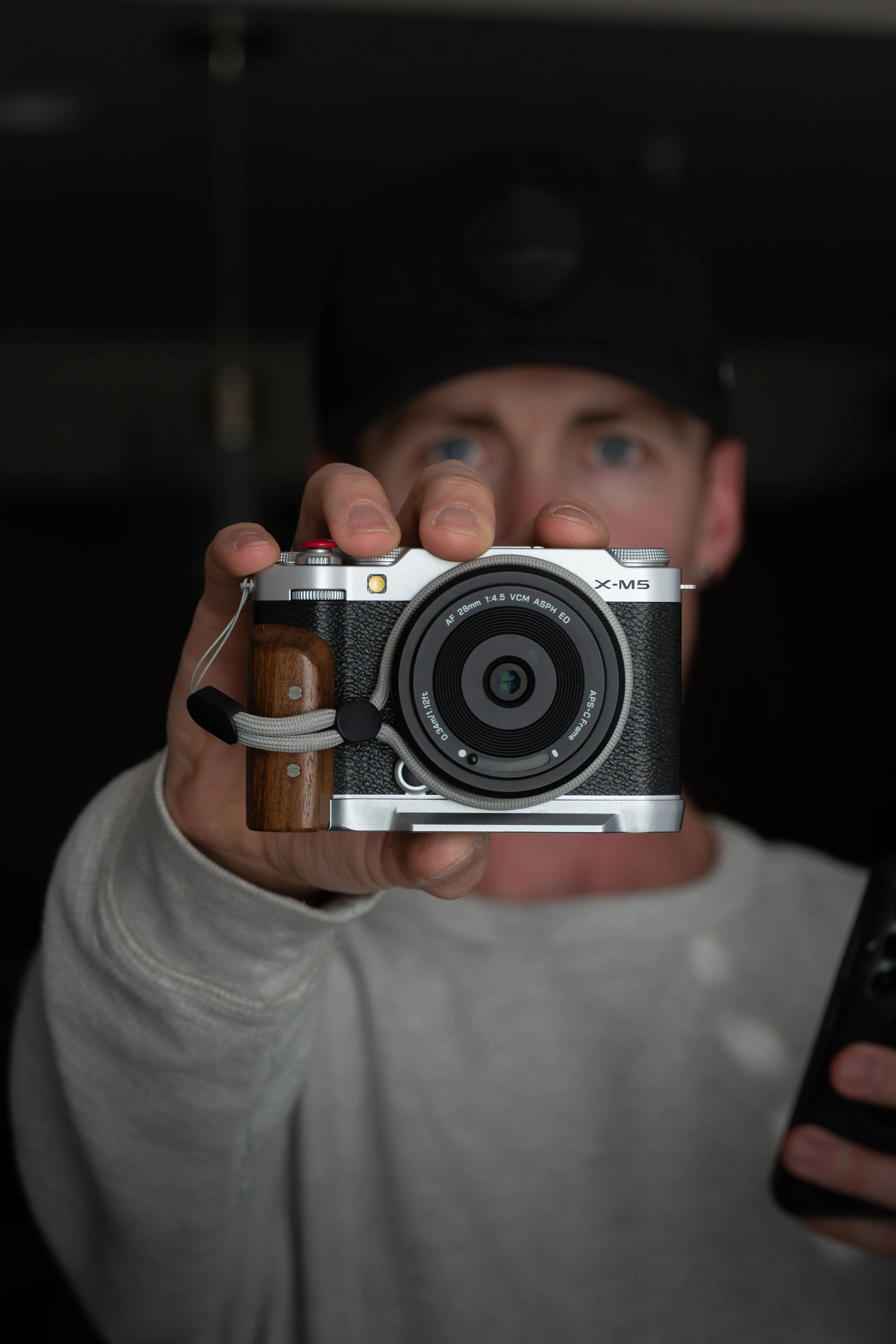 Person in gray sweater taking a photo with Fujifilm X-M5 camera, holding it with their right hand, wears a black cap with a headlamp, background is dark