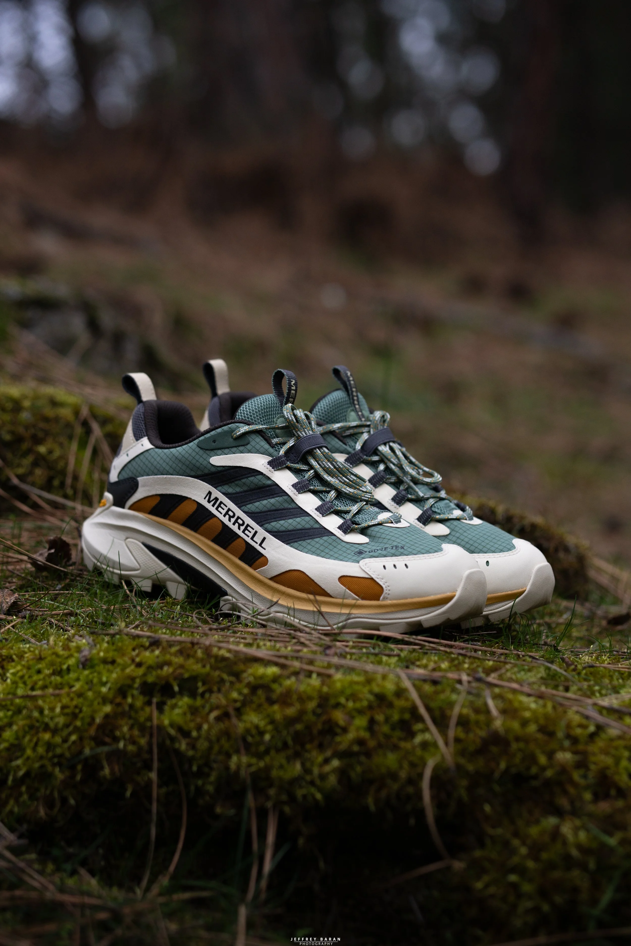 A pair of green, black, and white Merrill hiking shoes resting on mossy ground in a forest
