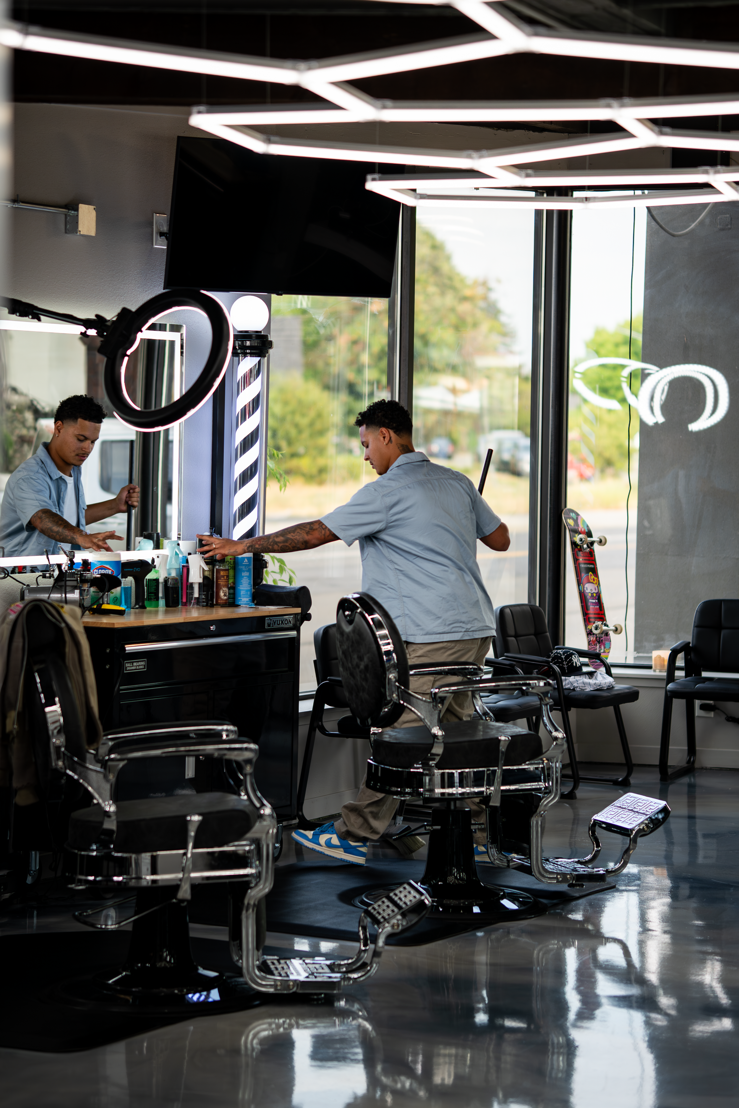Barbershop interior with two barbers working, barber chairs, hair-cutting supplies on a counter, a skateboard, and large windows with outdoor view.