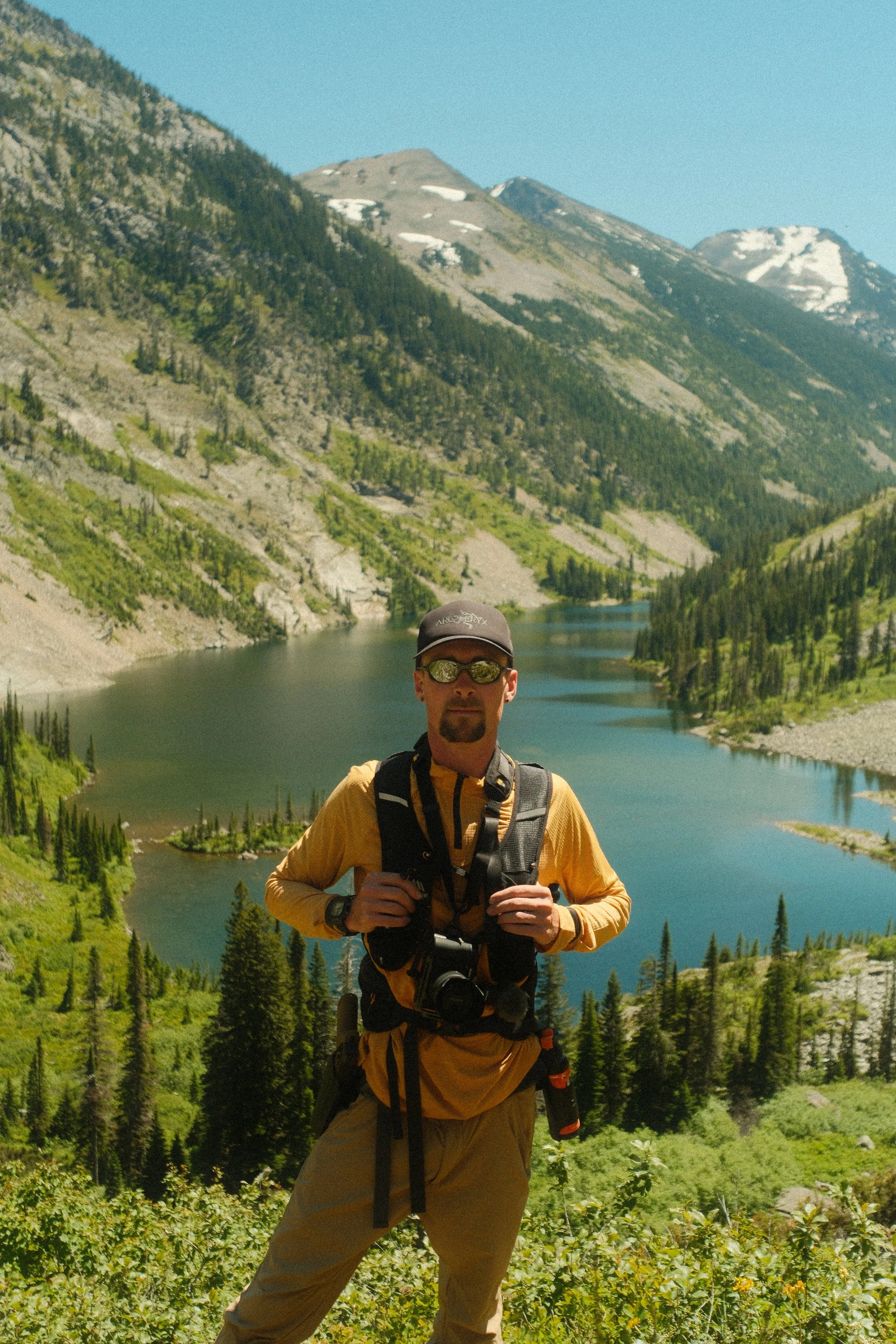 Man with sunglasses, cowboy hat, and yellow jacket standing in front of a river and mountains with snow patches, dense forest, and clear blue sky.