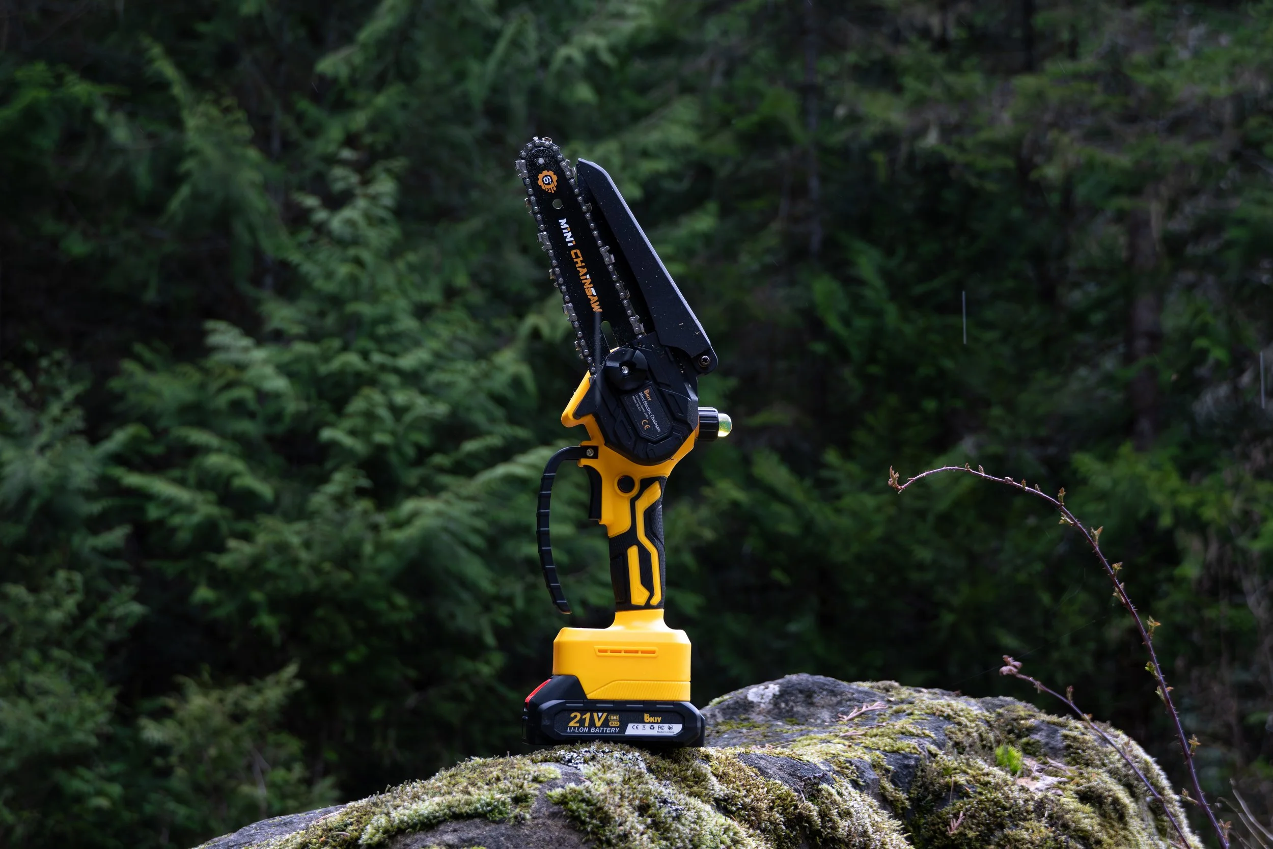 A yellow cordless chainsaw with black accents, placed on a moss-covered rock in a forest setting. The chainsaw has a blade with the brand name 'Mini Chainsaw' visible and a 21V lithium-ion battery attached at the bottom.