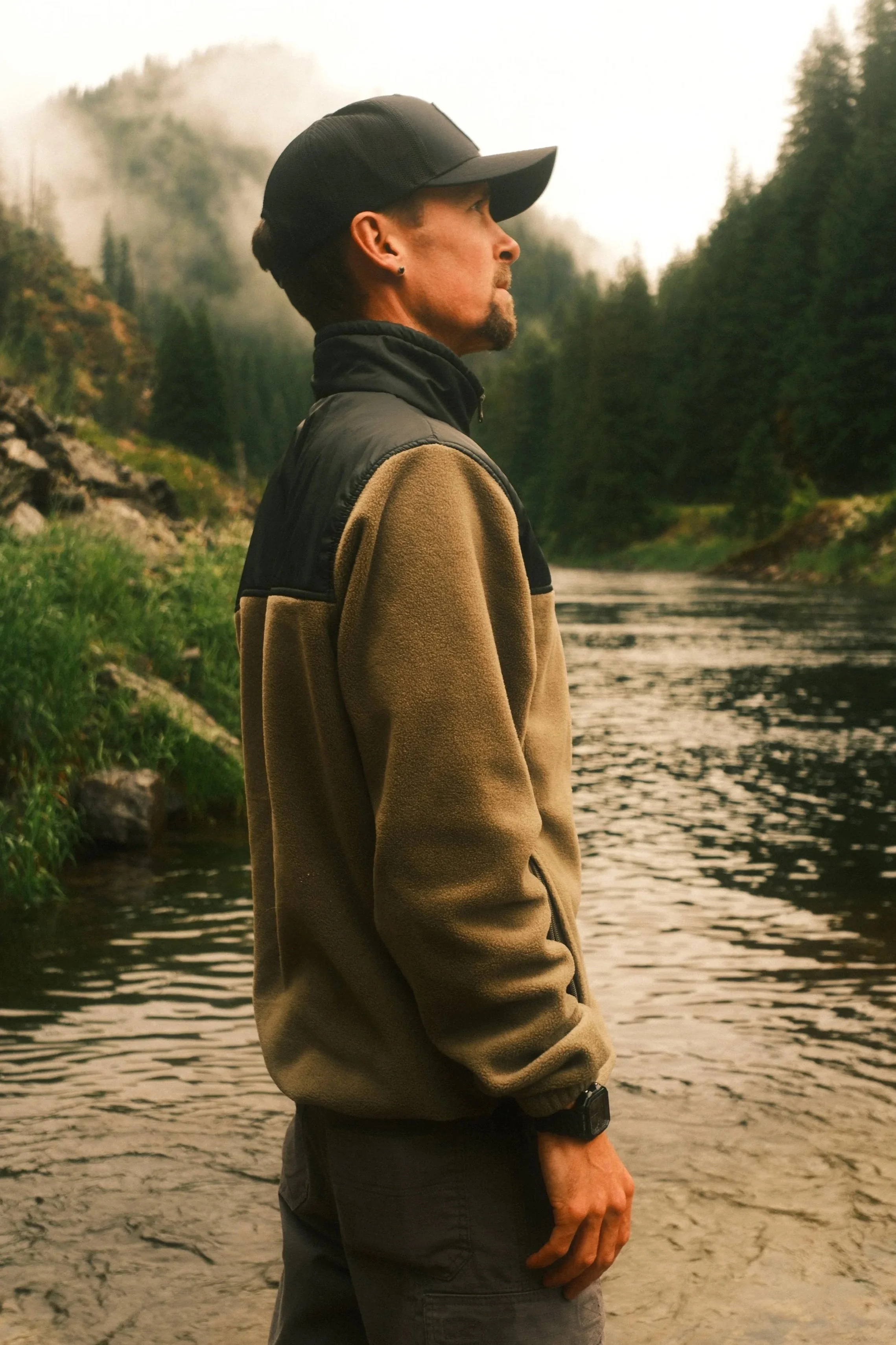 A man standing near a river, looking into the distance, surrounded by trees and mountains in a misty outdoor setting.