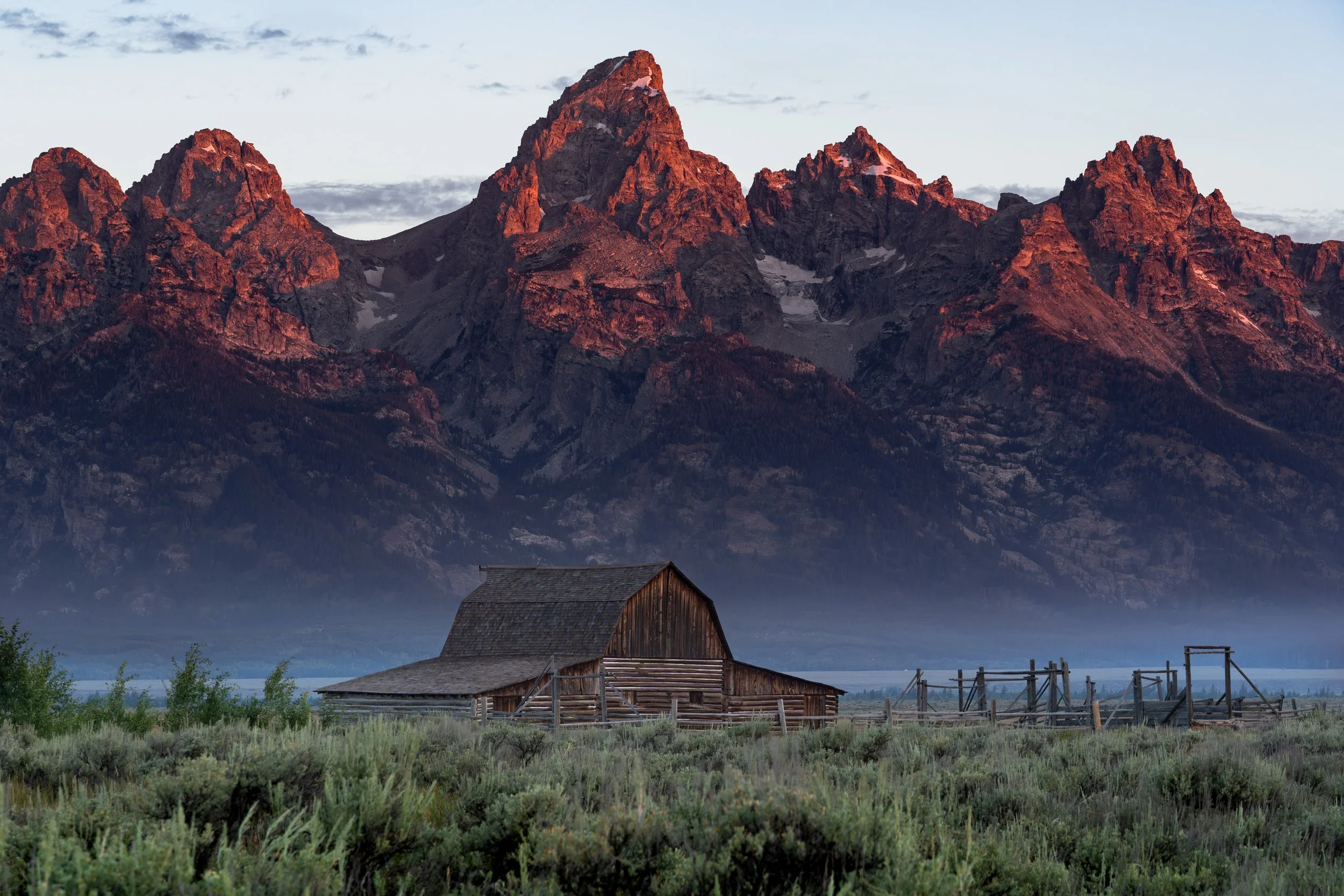 A rustic wooden barn in a grassy field with a mountain range in the background at sunset.