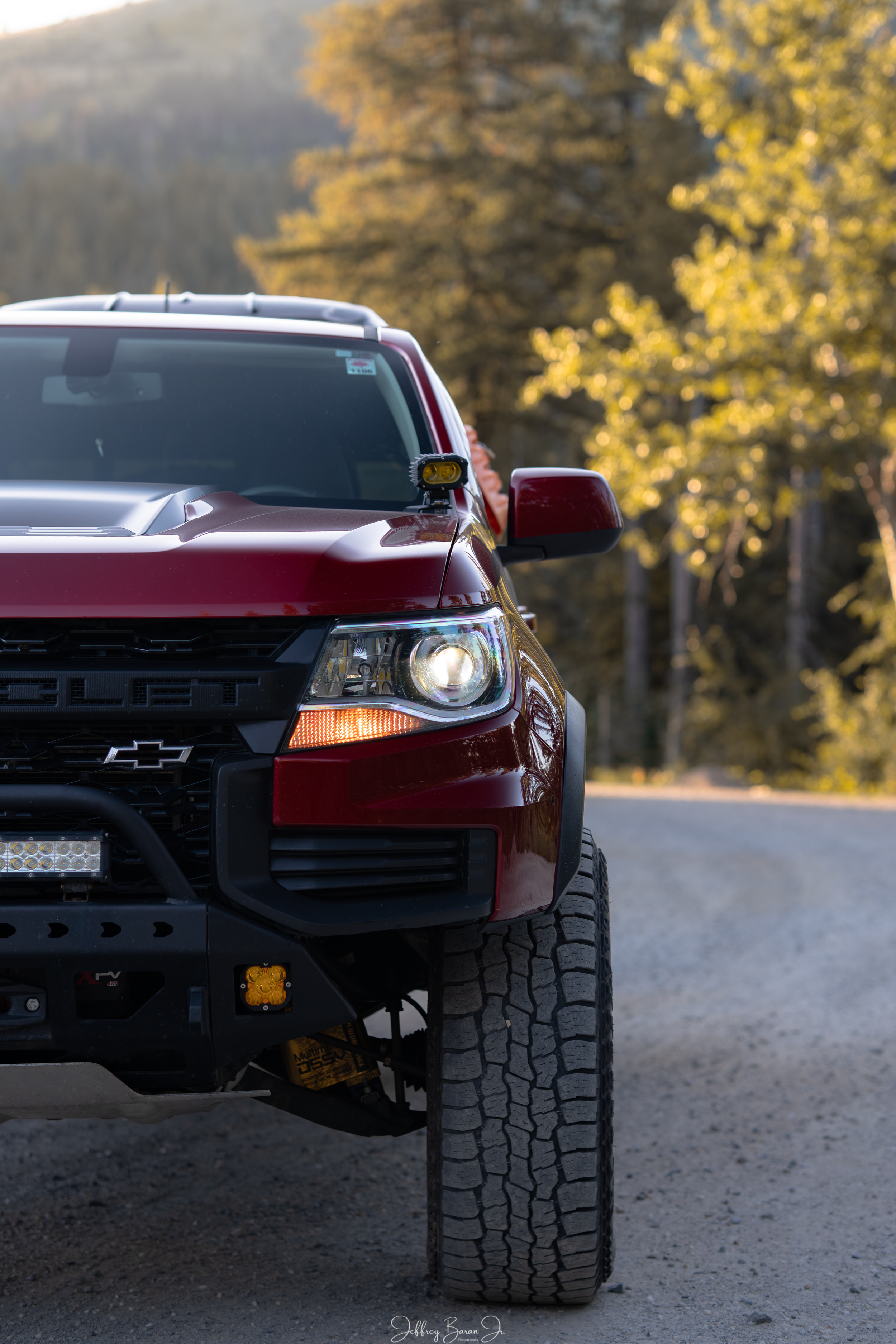 Close-up of the front part of a red Chevrolet pickup truck with large off-road tires, parking on a dirt road surrounded by trees with autumn foliage.