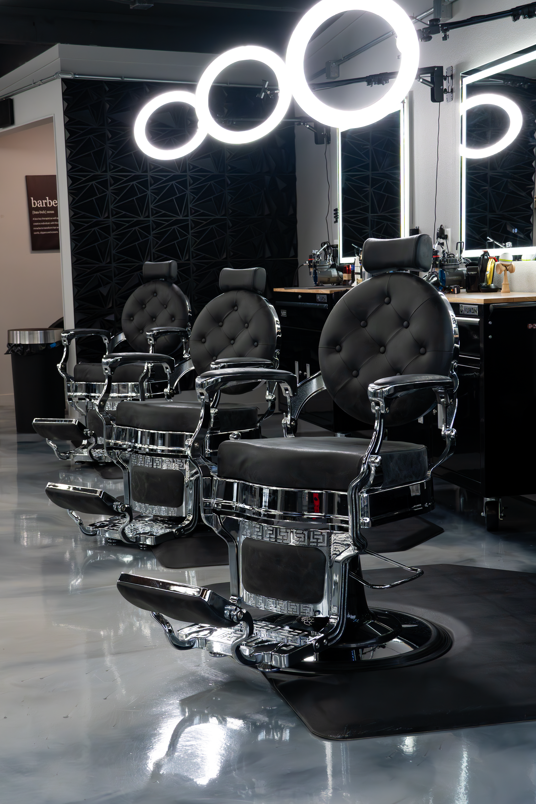 Barber chairs in a modern salon, illuminated mirror, black textured wall, and barber tools on the counter.