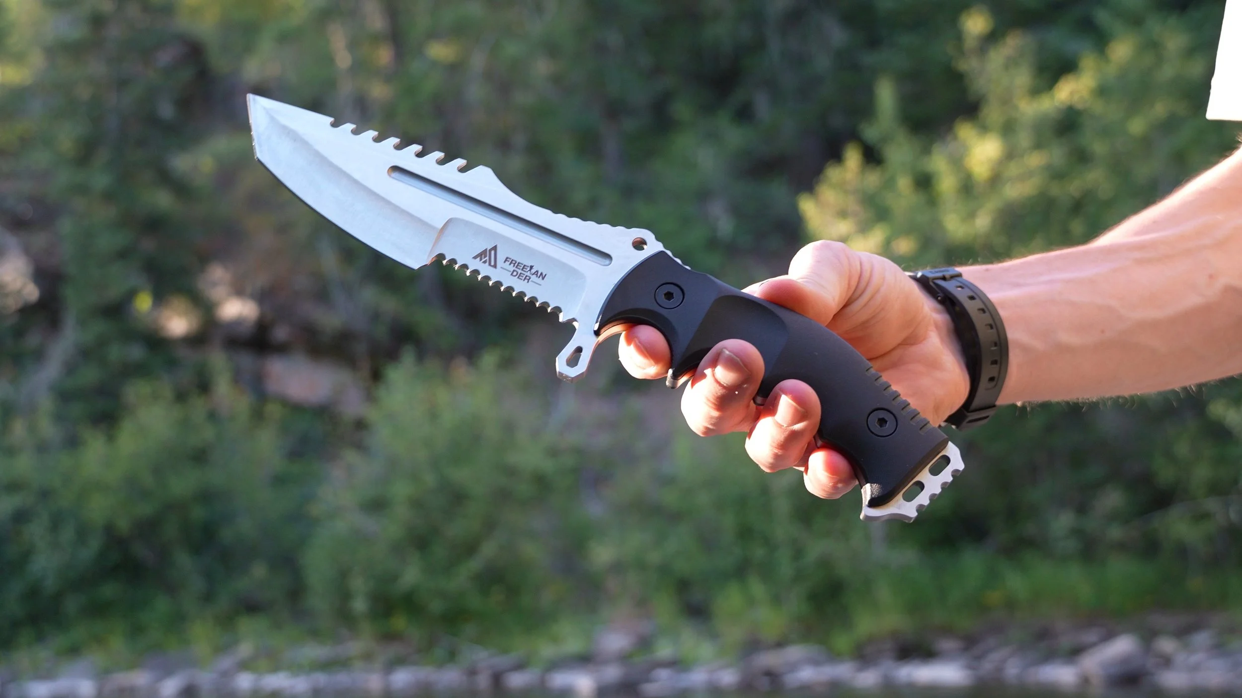 A person holding a tactical knife with a serrated blade, black handle, and metal accents outdoors with blurred trees in the background.