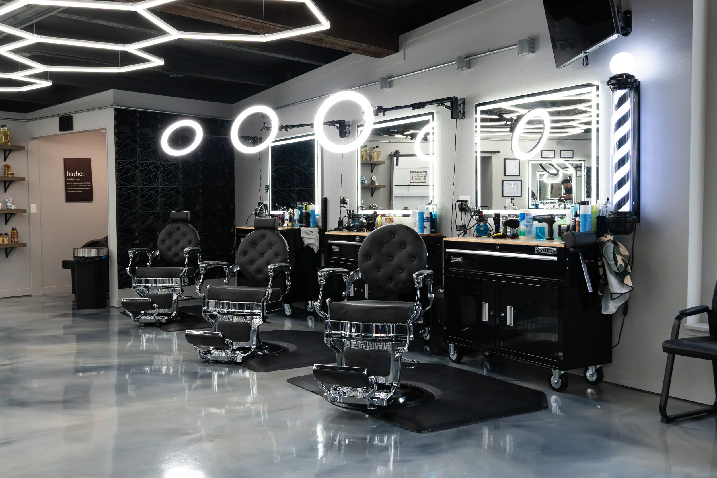A modern barbershop with multiple black chairs in front of mirrors with bright circular LED lights, a black work station with barber tools and supplies, and a white wall with shelves holding hair care products.