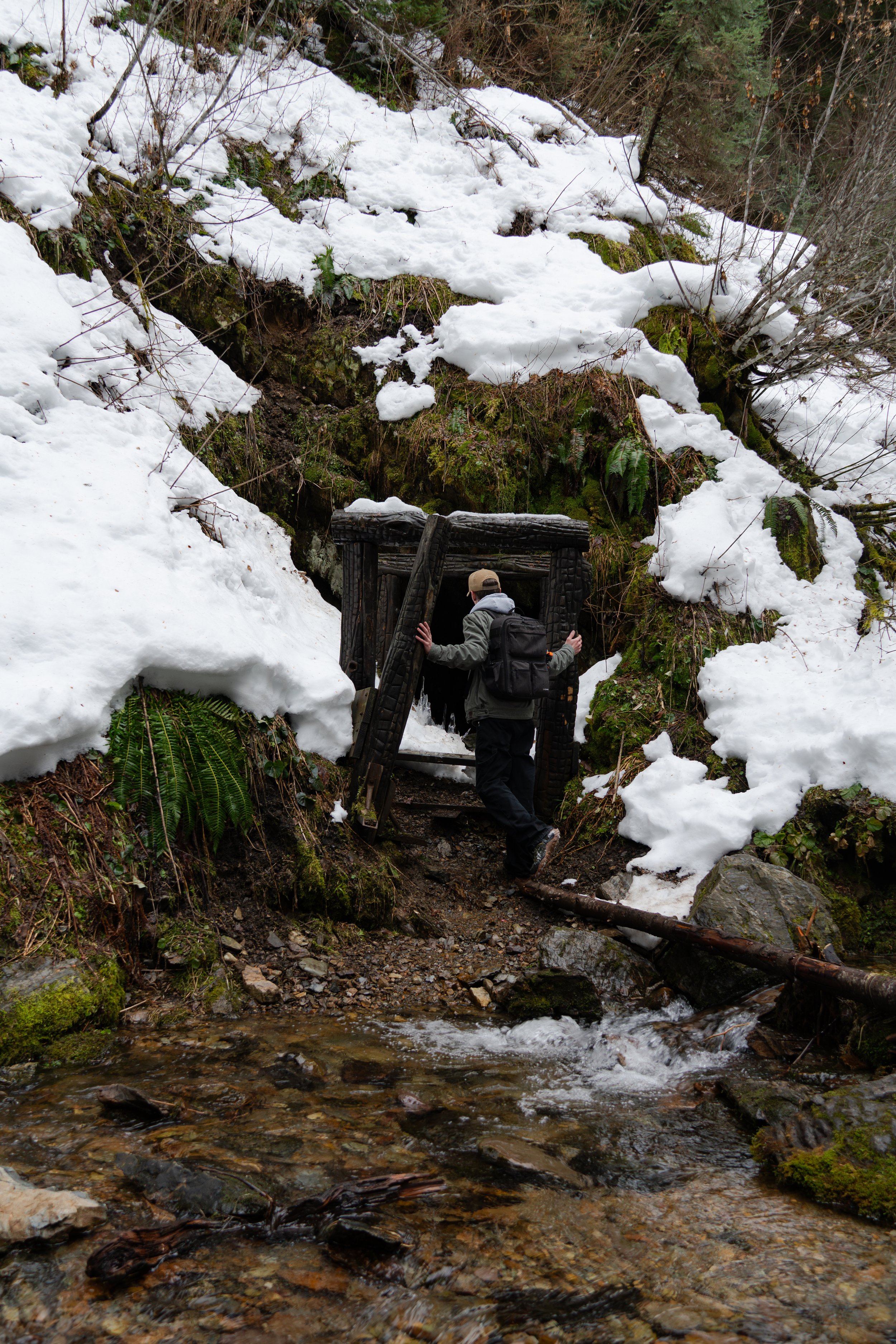 A person in outdoor gear, including a backpack and hat, stands at a large, wooden, framed doorway on a snowy hillside with a small stream flowing at their feet and snow-covered rocks and trees in the background.