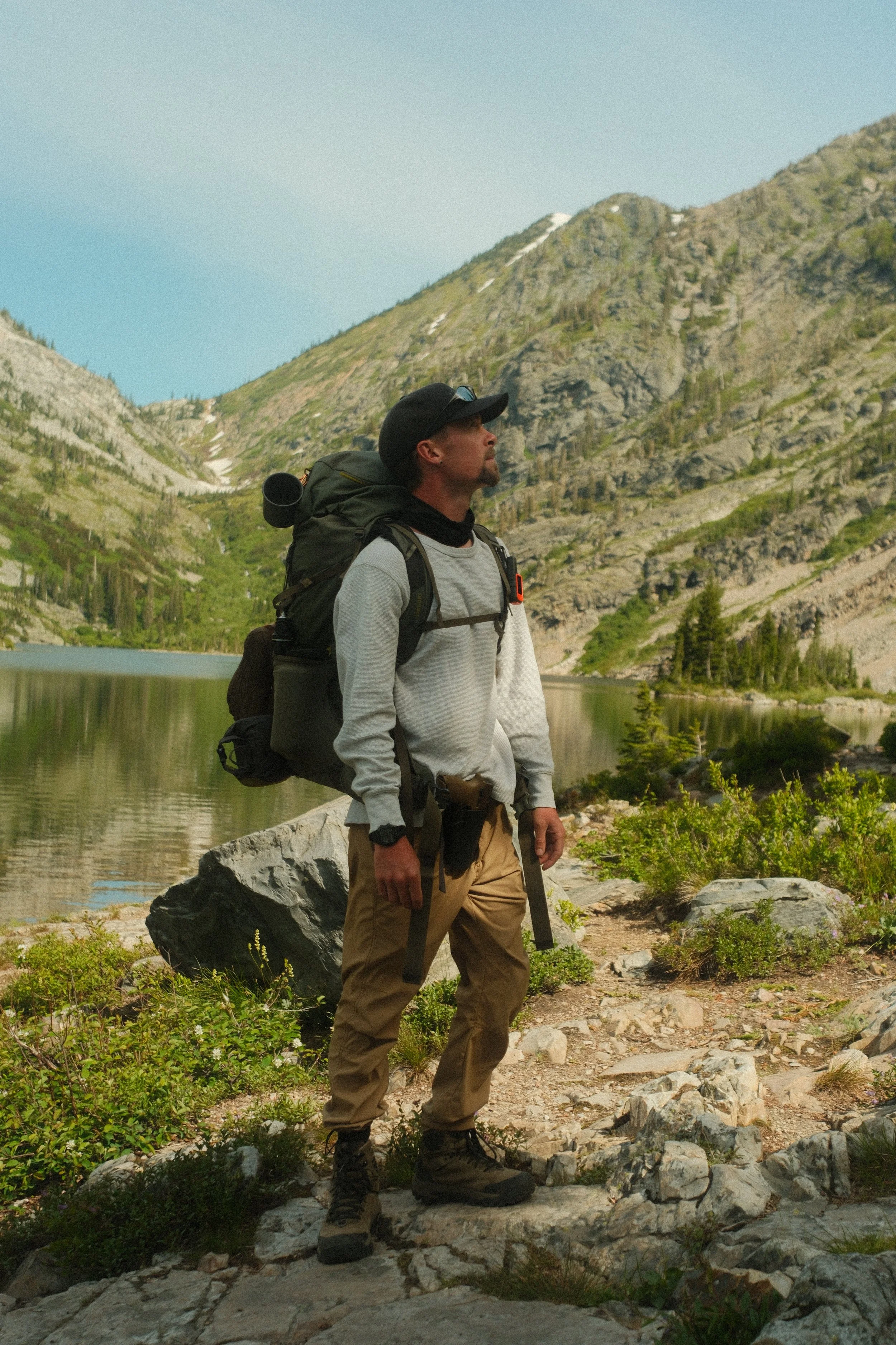 A man with a beard wearing a gray sweatshirt, tan cargo pants, and hiking boots stands on rocky terrain near a lake with mountains and greenery in the background. He has a black backpack and a black cap.