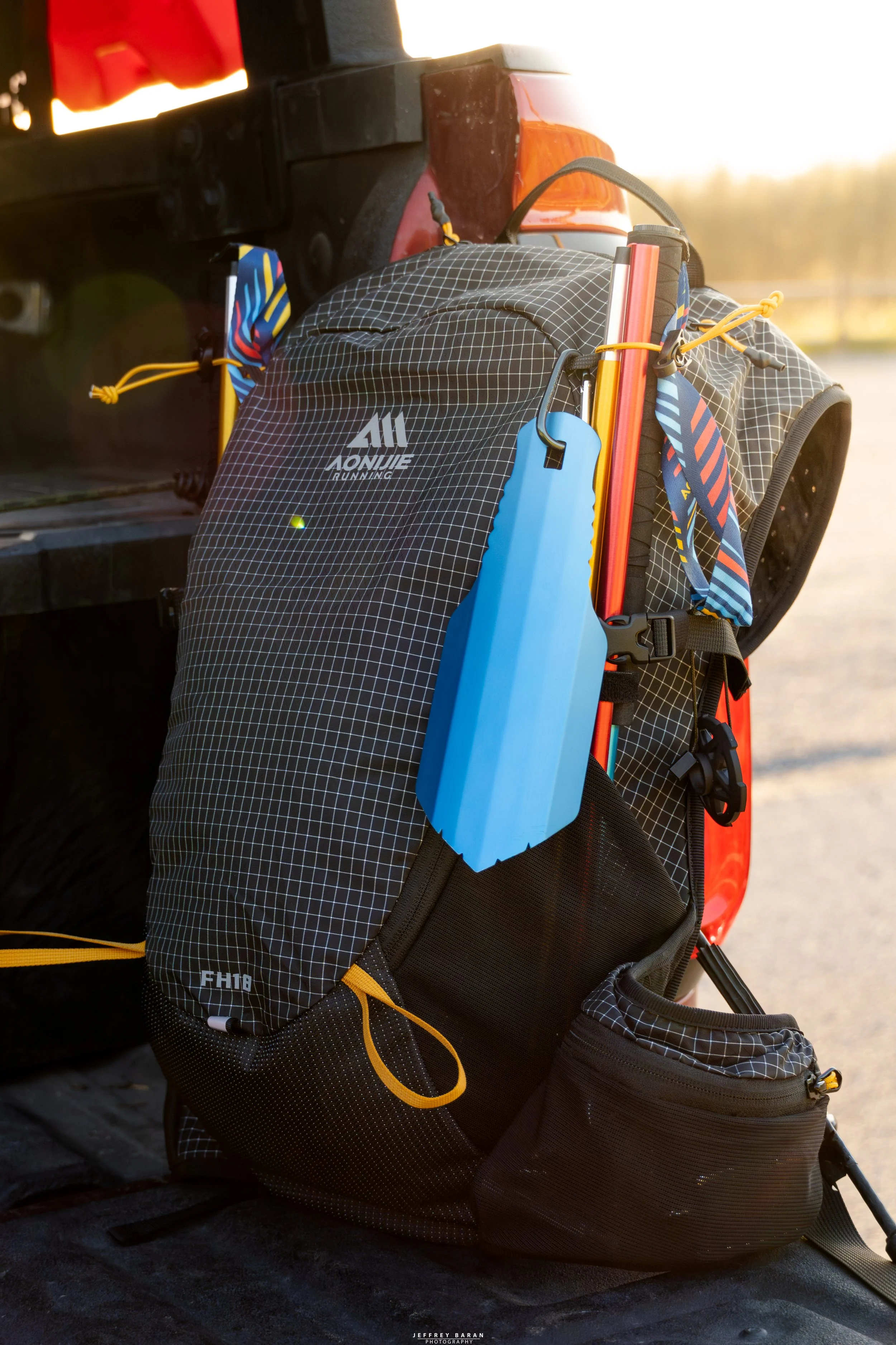 A black and gray Osprey hiking backpack with yellow accents, placed on a black surface outdoors with a blurred background of trees and sky, in warm sunlight.