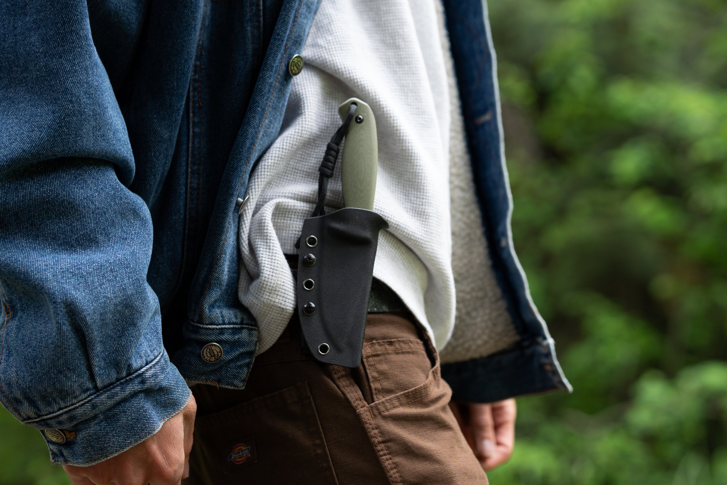 Close-up of three people standing outdoors, wearing layered clothing including denim and fleece, with a small outdoor pocket knife clipped to their clothing.