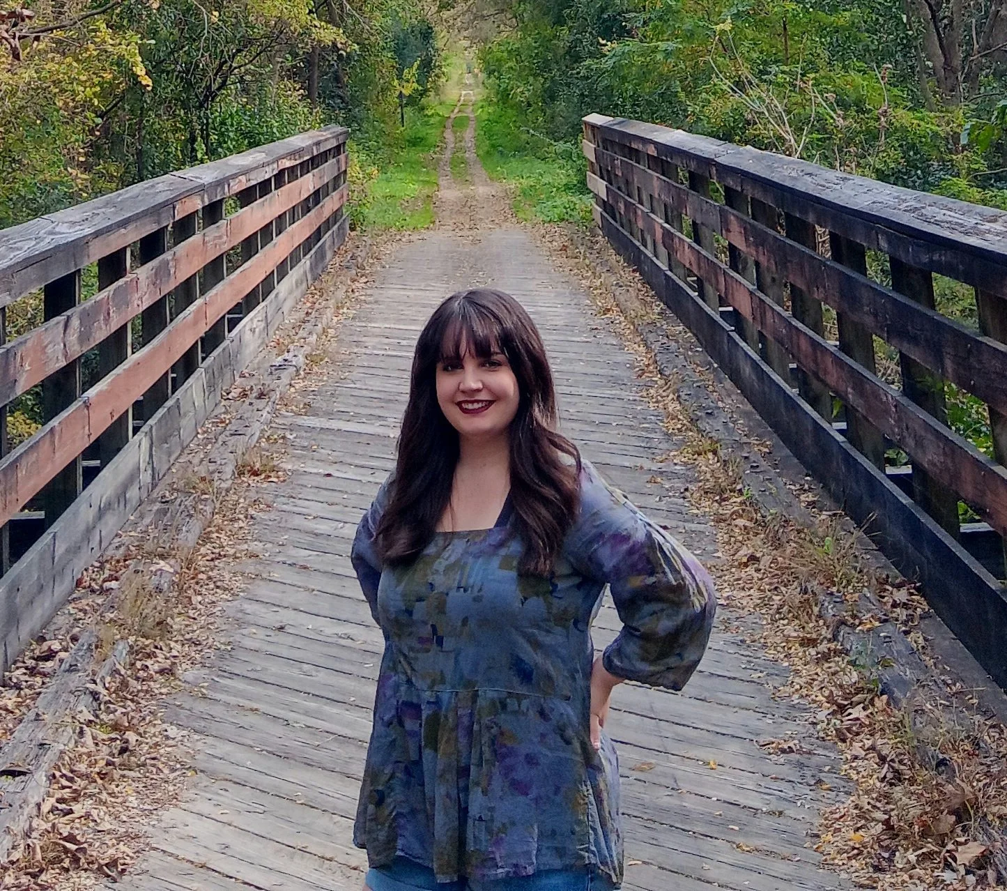 A woman with dark hair and a patterned dress standing on a wooden bridge surrounded by trees.