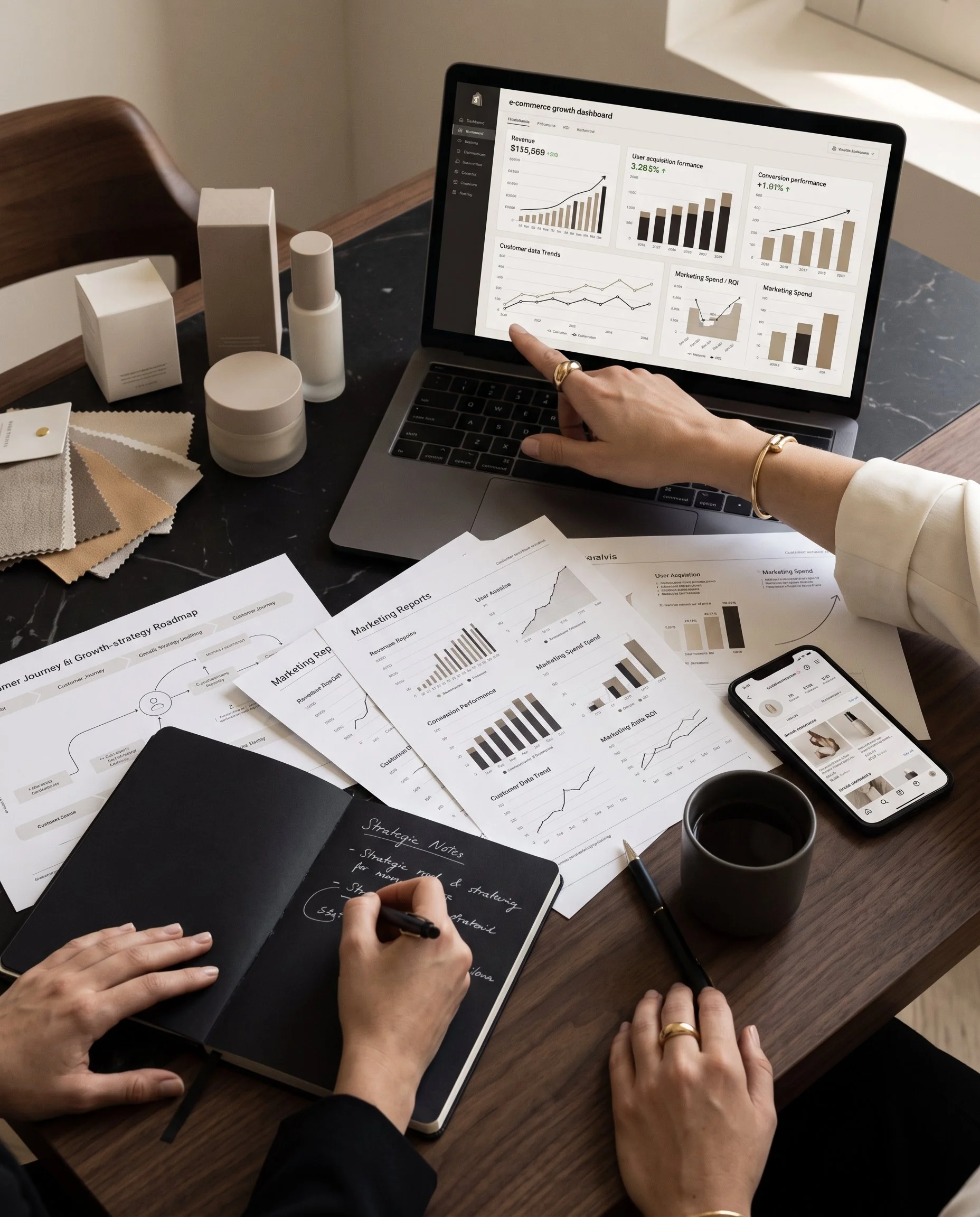A person writing in a black notebook amid scattered papers, charts, and graphs on a wooden table, with a laptop displaying an e-commerce dashboard, a smartphone, a pen, and a coffee mug nearby.