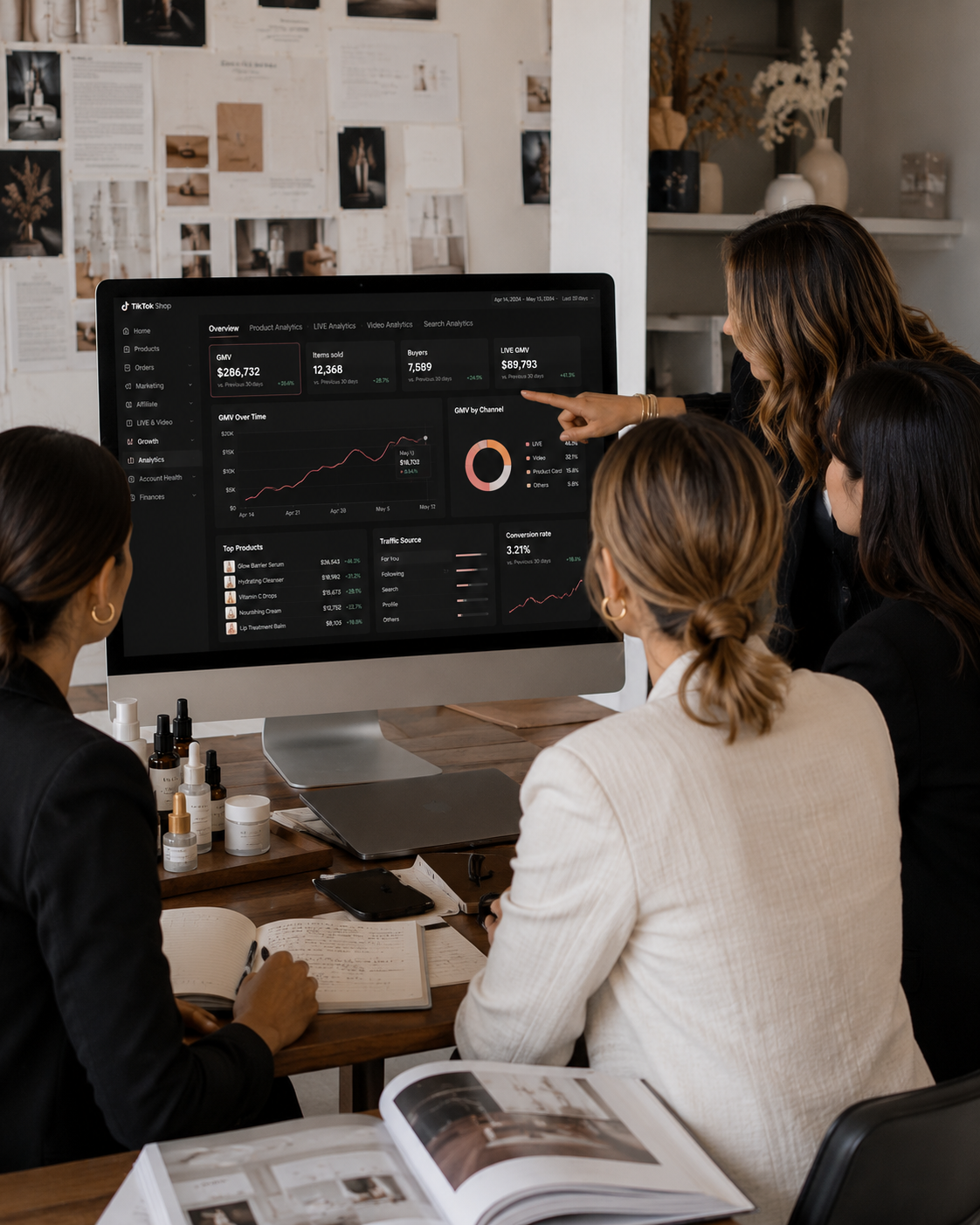 Four women in business attire gathered around a desk, analyzing data on a large computer monitor with various charts and graphs in a modern office setting.