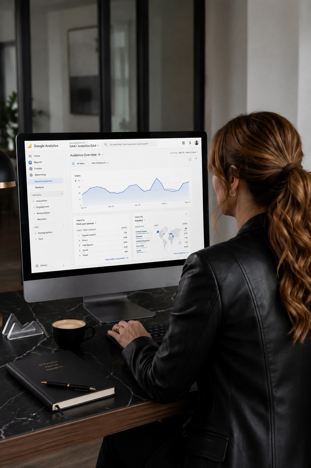 A woman with brown hair tied back, wearing a black leather jacket, working on a computer displaying Google Analytics data in a modern office.