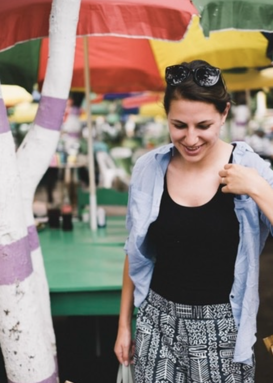 Smiling woman wearing sunglasses and patterned pants at an outdoor market beneath colorful umbrellas