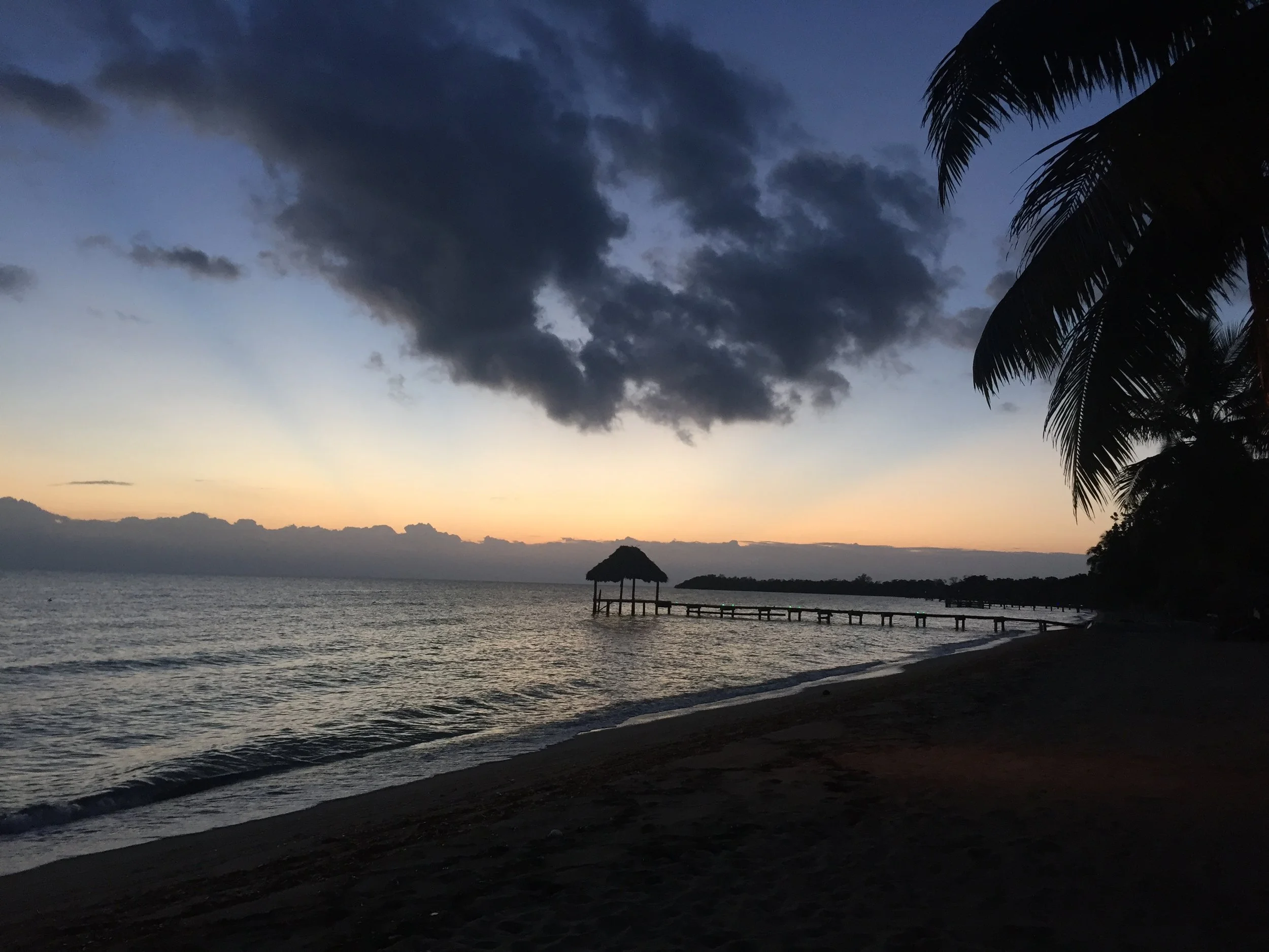 Tropical beach at sunset with a thatched-roof pier, calm ocean, palm trees, and dramatic clouds