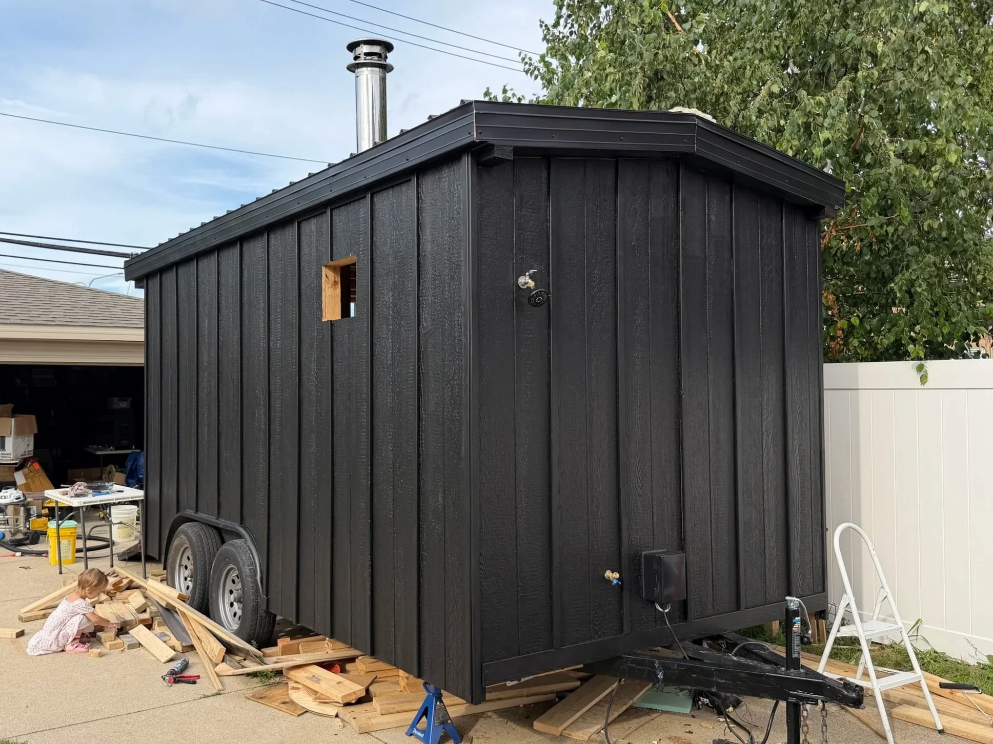 Black tiny house on wheels in driveway, children playing with wood, tools, and construction materials, large tree in background, house in progress.