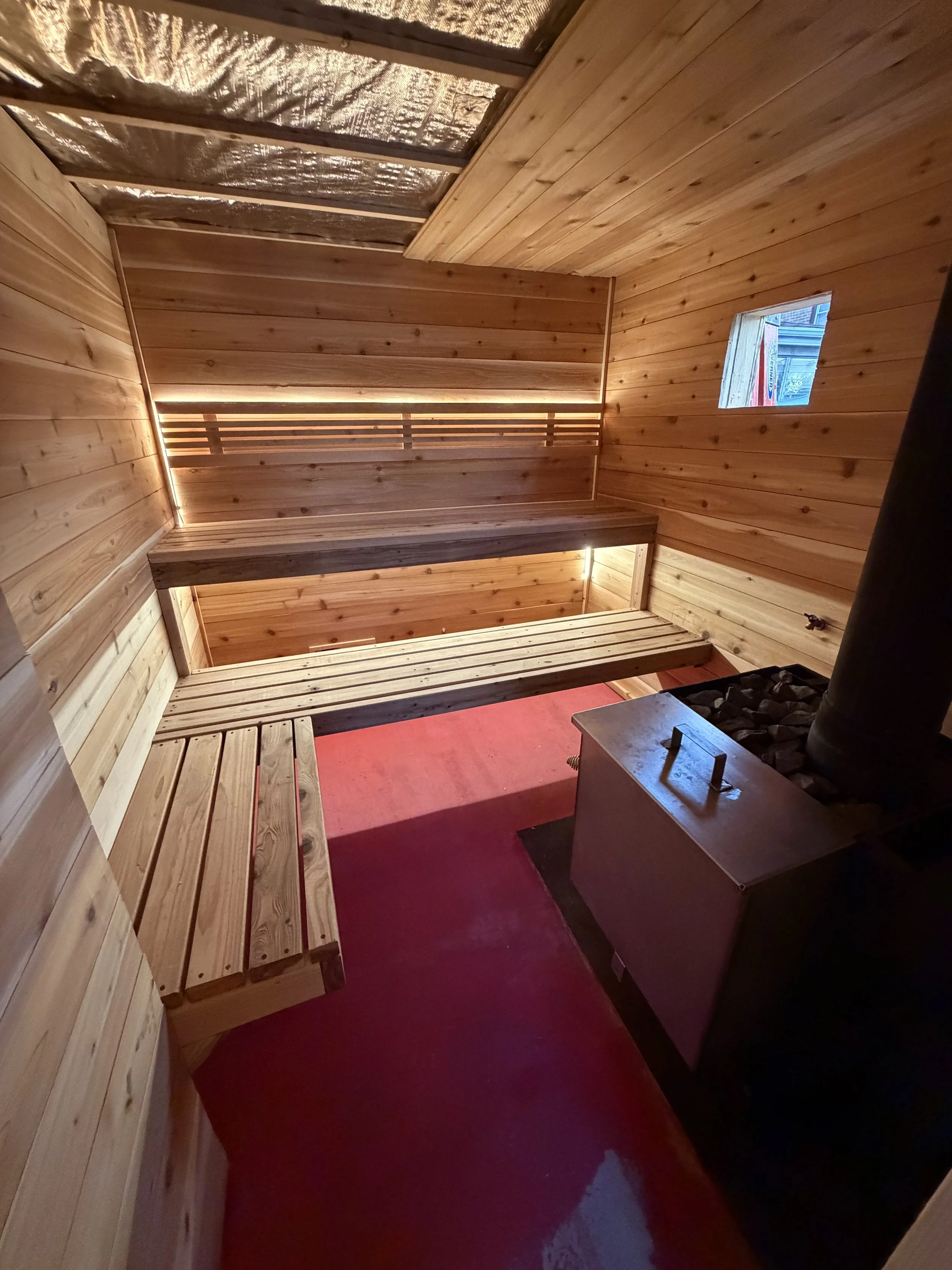 Interior of a wooden sauna with benches, a small window, and a wood stove with stones.