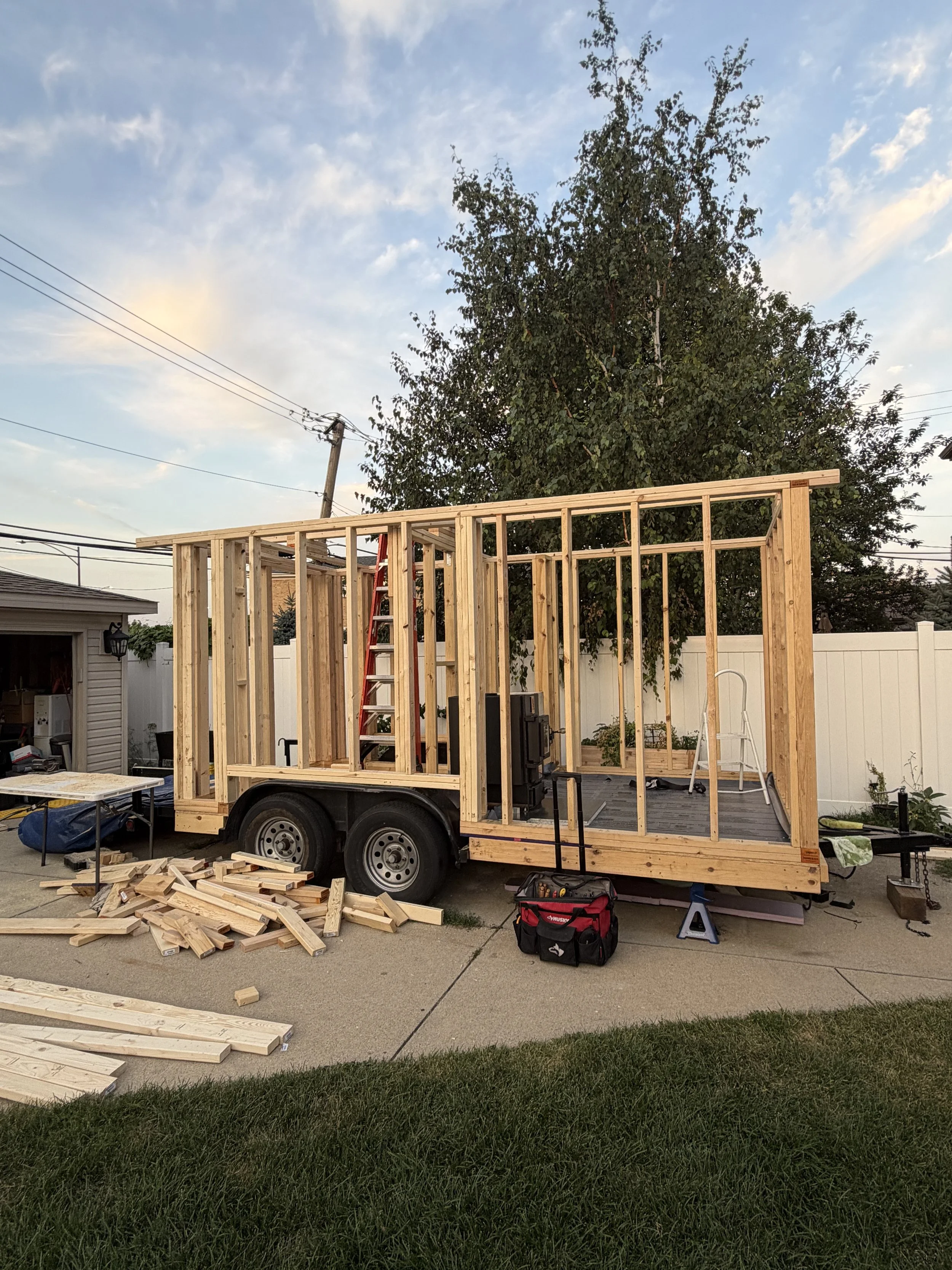 Wooden framing of a small structure built on a trailer in a backyard with tools and materials nearby, under a partly cloudy sky.
