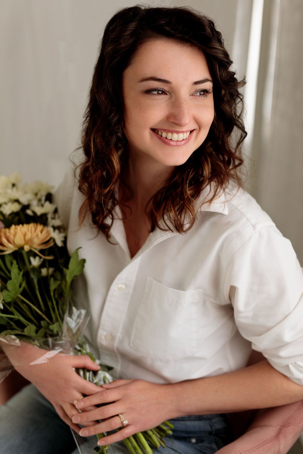 A woman with curly brown hair, smiling, holding a bouquet of flowers, wearing a white shirt, seated indoors.
