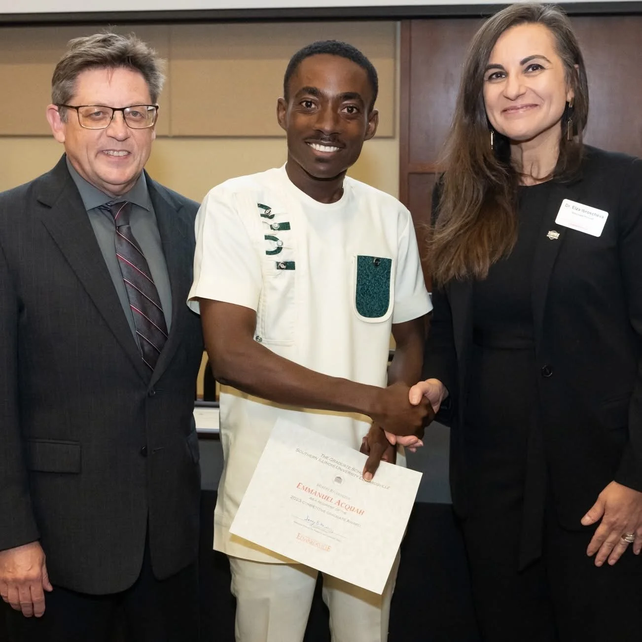 A man in a white medical uniform shaking hands with a woman in a black blazer, while another man in a suit stands beside them during an award or certification ceremony.
