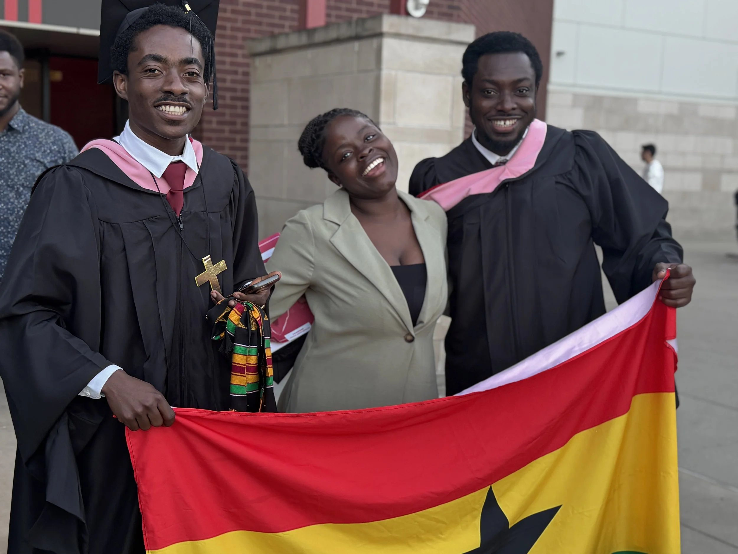 Three smiling individuals, two men and one woman, holding a Ugandan flag during a celebration or graduation event outside a brick building.