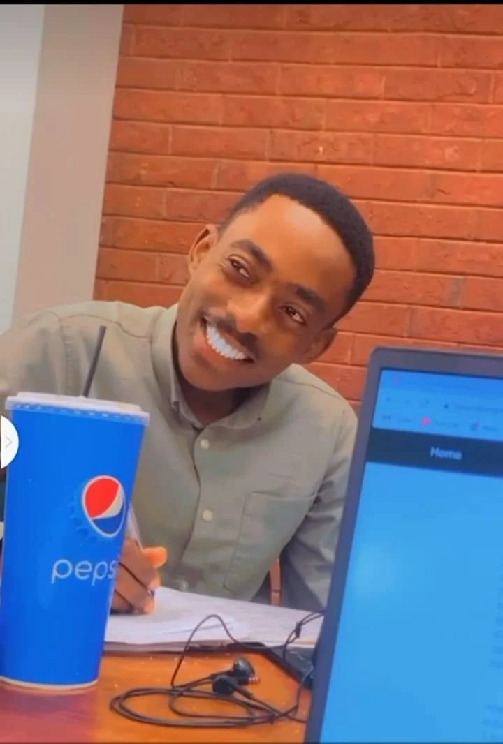 A man smiling at a table with a Pepsi cup, a notebook, and earphones, in front of a laptop and a brick wall background.