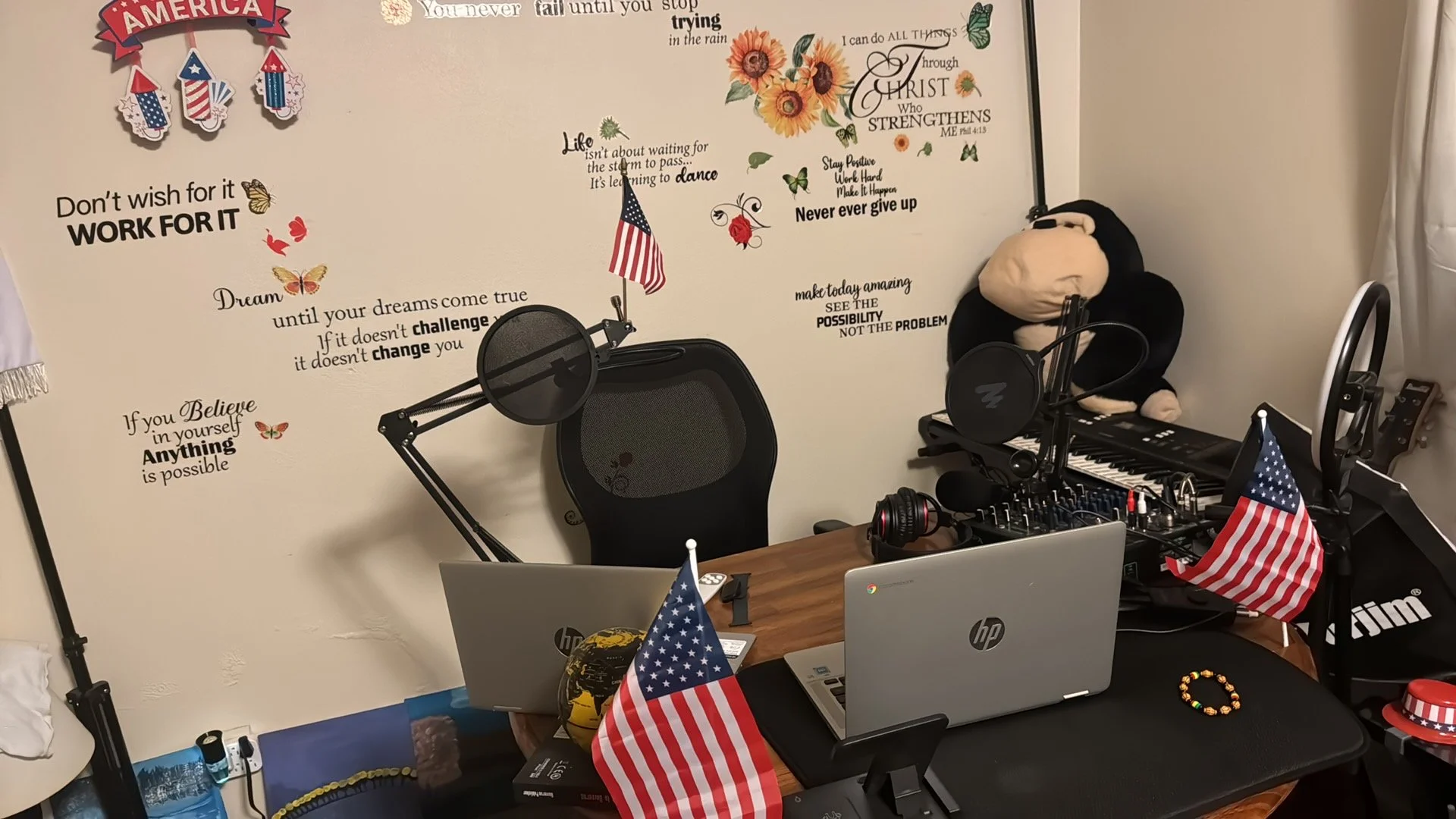 A home office decorated with American-themed items, including multiple small American flags, a large wall sticker with motivational quotes and sunflower graphics, a plush monkey toy on a keyboard stand, two laptops on a wooden desk, microphones, a pair of headphones, and other electronic equipment.
