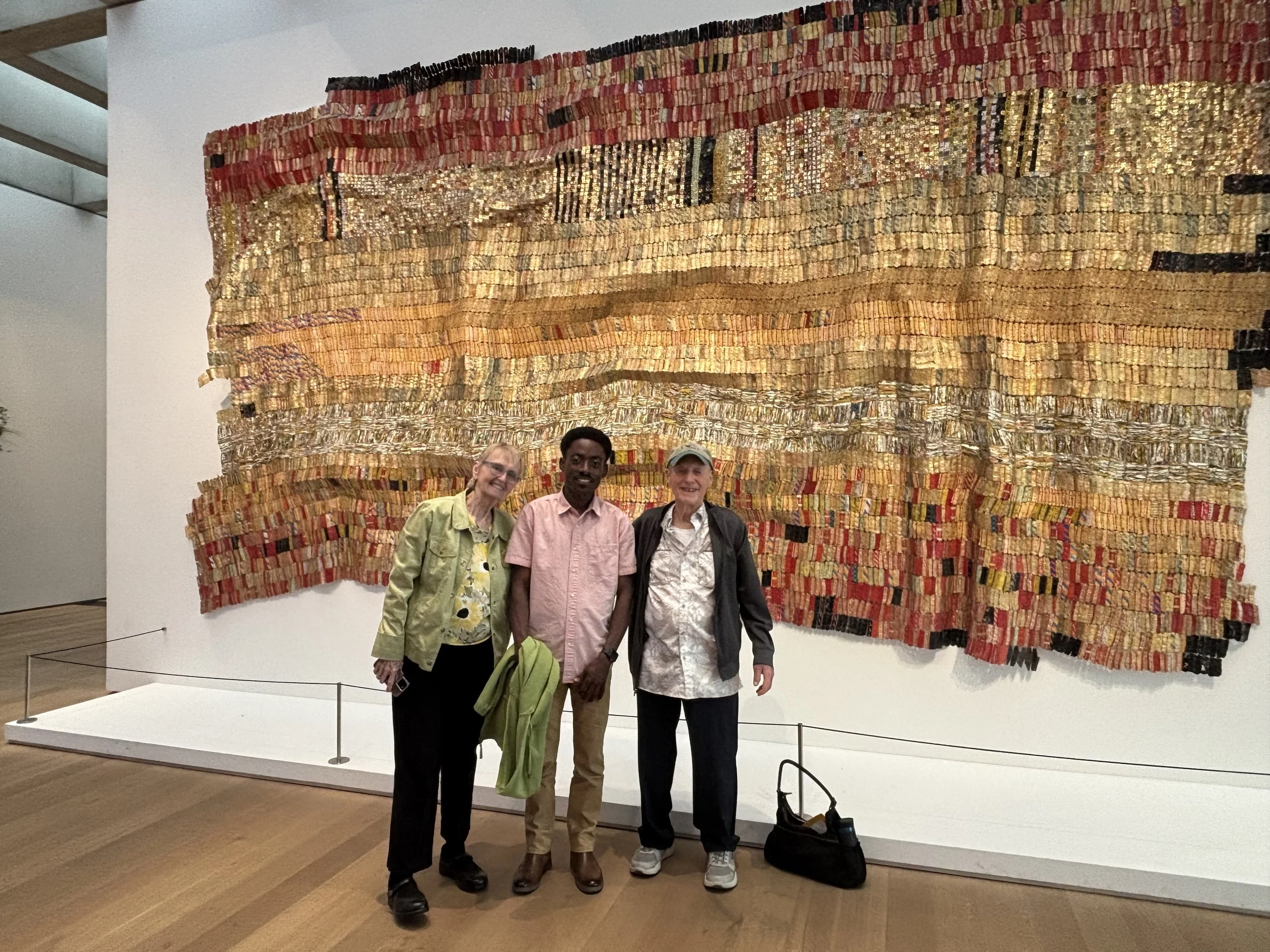 Three people standing in front of an abstract woven artwork in a museum. They are smiling and posing for the photo.