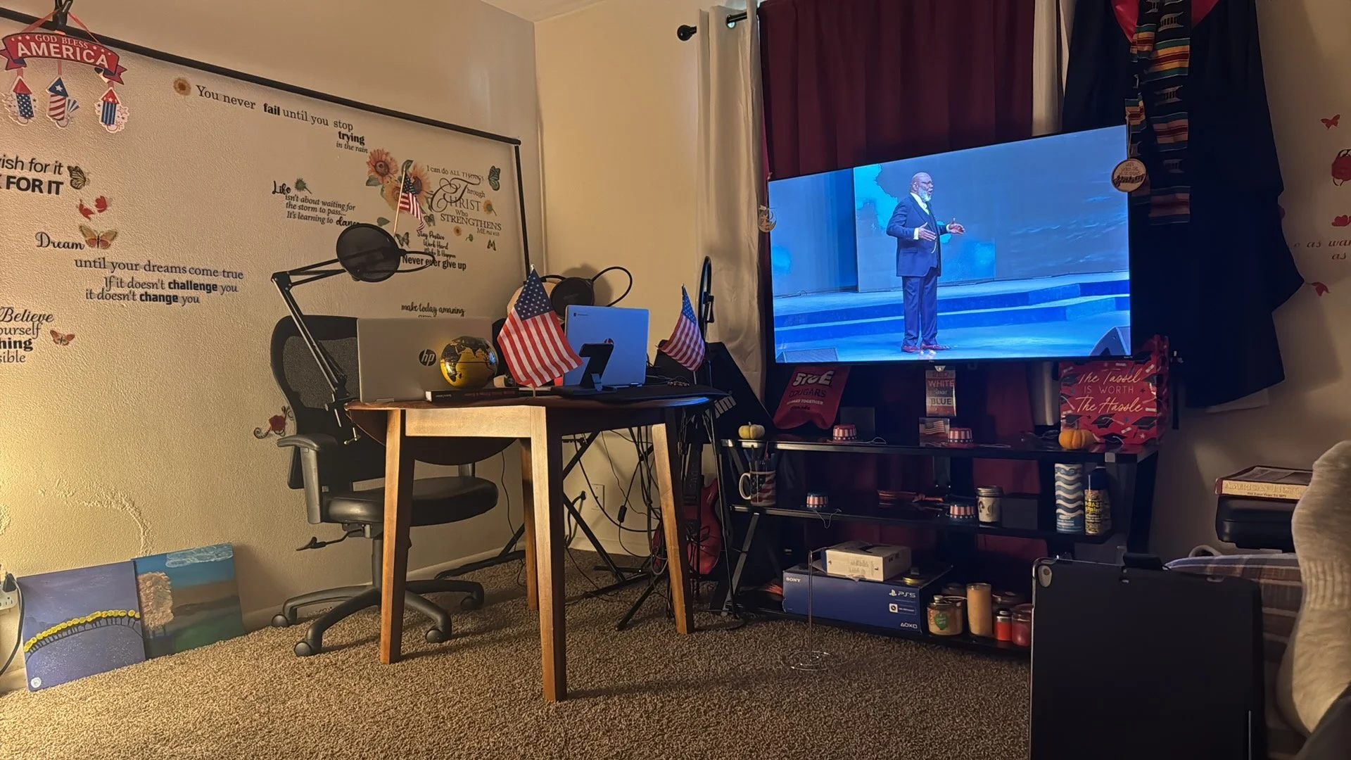 A living room with a large flat-screen TV showing a man in a suit giving a speech on stage. The room has red curtains, an off-white wall with inspirational quotes and a decorative quote about America. There are two small American flags on a desk with a computer and headphones. There are some paintings on the carpeted floor and various items including mugs, a box, and candles on a black shelving unit and a table.