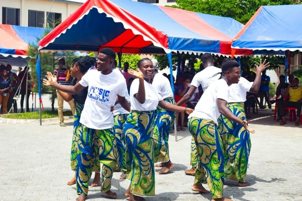 Young people dancing outdoors at a cultural event, wearing colorful skirts and white shirts, under tents with red, blue, and orange patterns.