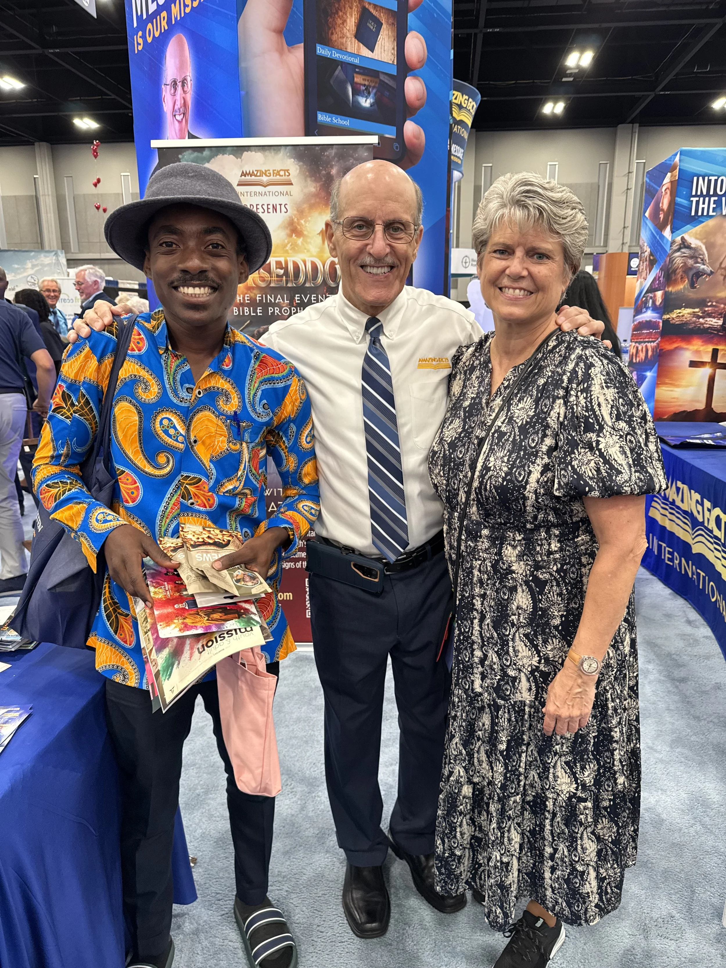 Three people standing close together at a conference or event, smiling at the camera. The person on the left is wearing a colorful patterned shirt and a gray hat. The person in the middle is wearing glasses, a white shirt with a striped tie, and has their arm around the person on the left. The person on the right has short gray hair, a patterned dress, and is also smiling. The background features banners promoting religious or inspirational content, with one banner showing a hand holding a phone with devotionals and Bible studies.