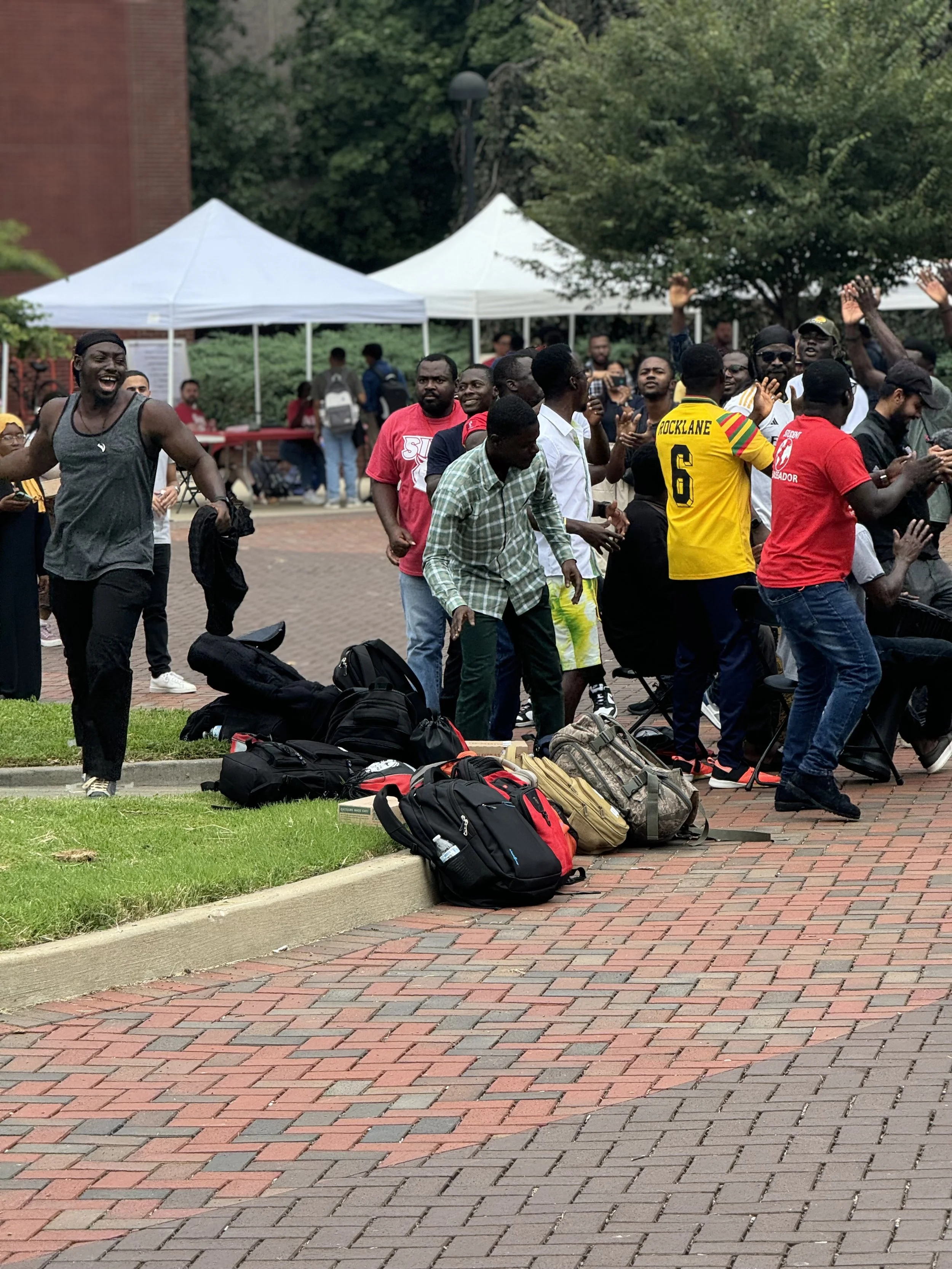 A group of international students, some sitting and some standing, with backpacks on the ground. Tents are set up in the background, and there are trees and a brick building nearby. The people appear to be engaged in a social or community event.