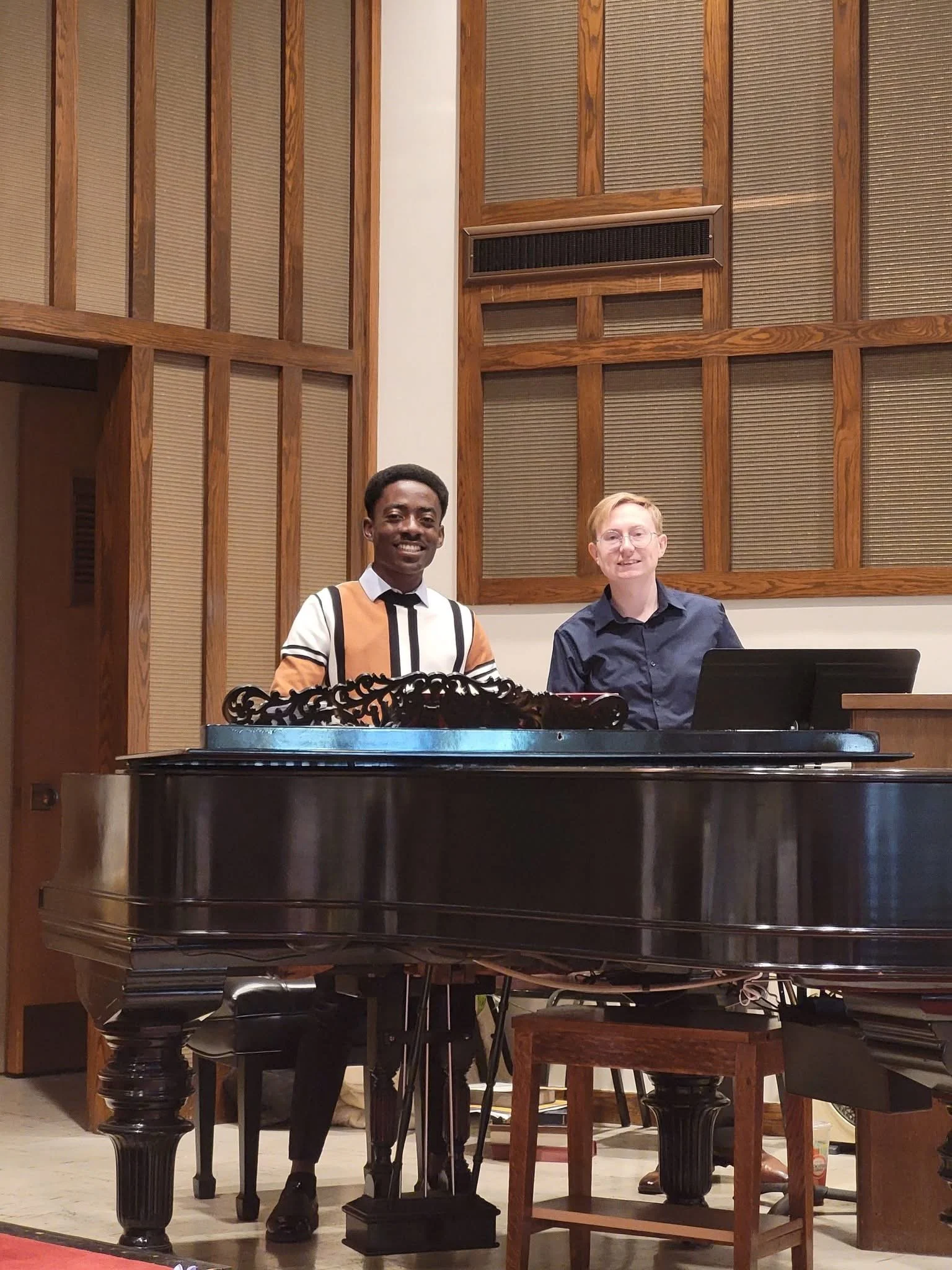 Two people, one with dark skin and the other with light skin, sitting at a black grand piano in a room with wooden wall panels.