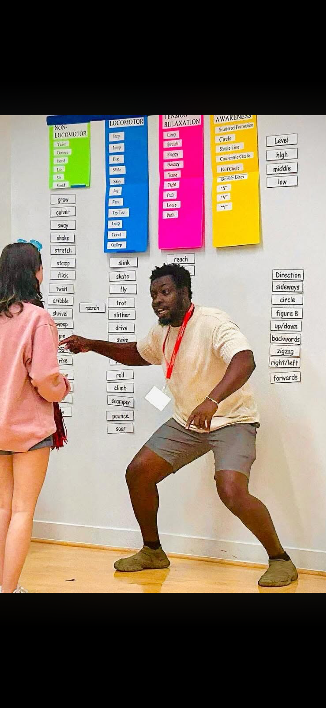 Two people, a man and a woman, are standing in front of a whiteboard with colorful charts and typed words. The man is wearing a beige shirt and shorts, and appears to be explaining or presenting something animatedly, pointing towards the whiteboard.