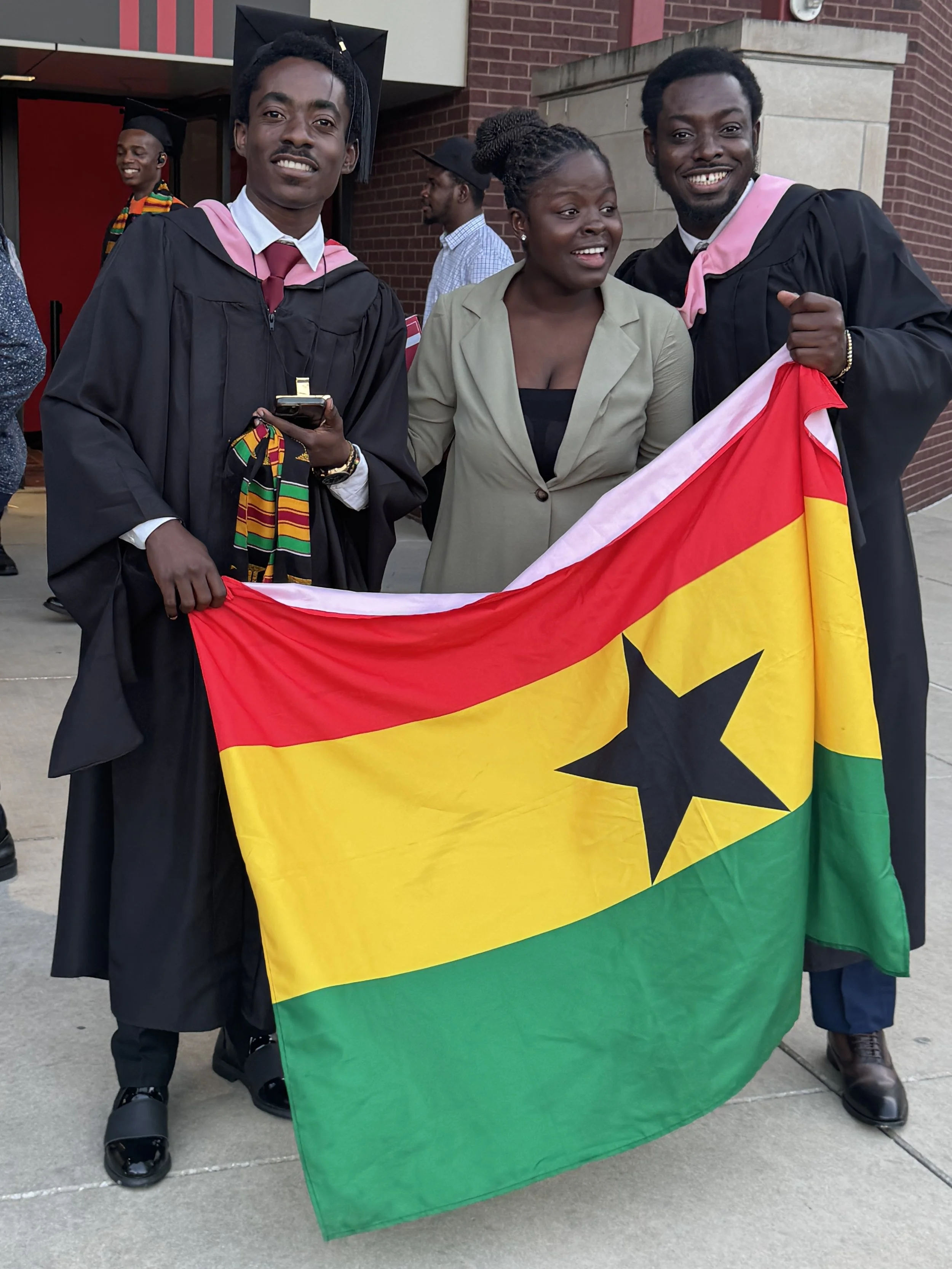 Group of three graduates including Prophakwa celebrating graduation outside, holding a Ghanaian flag, wearing caps and gowns, with a brick building in the background.
