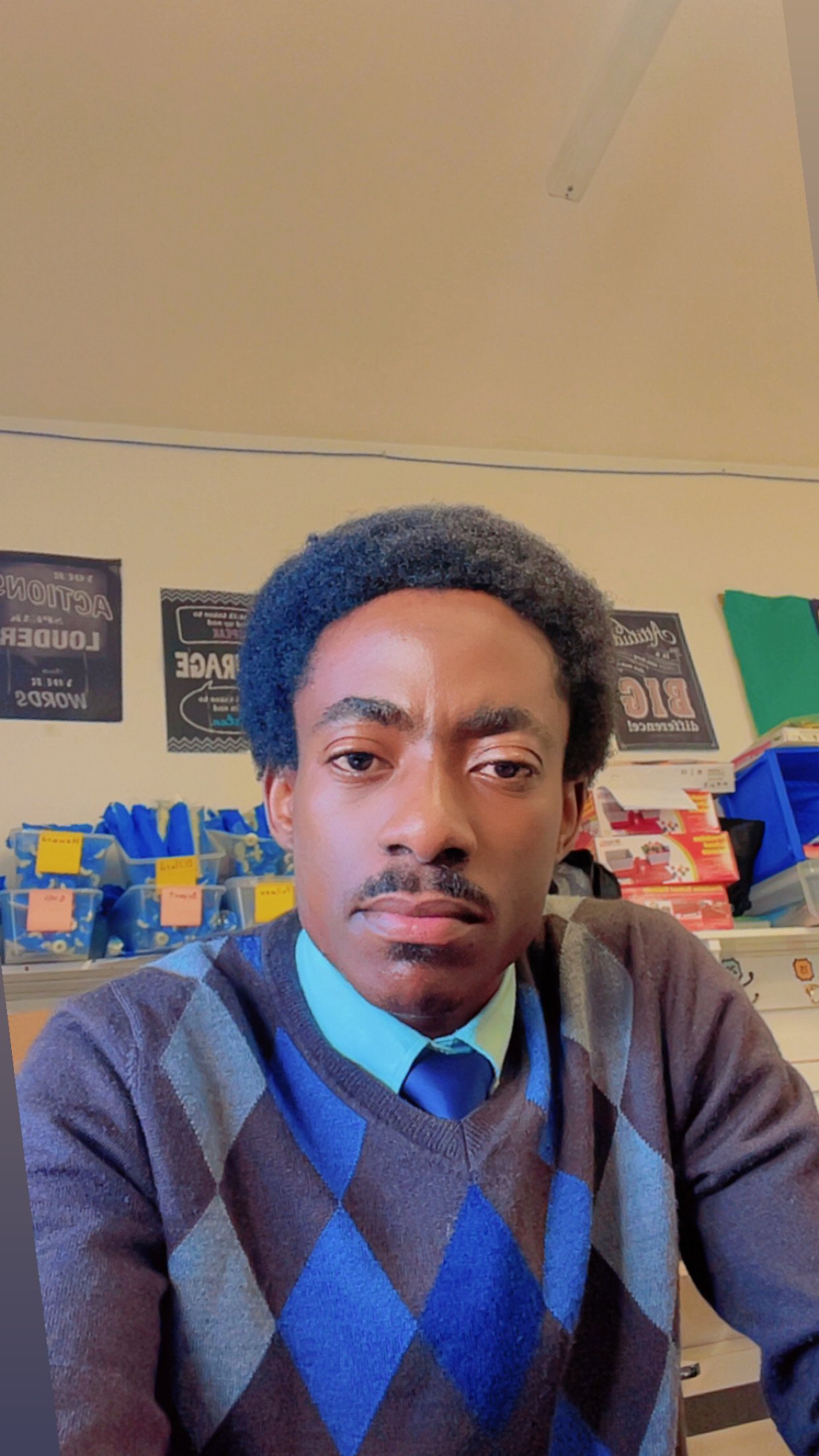 A young man with dark curly hair, wearing a blue argyle sweater vest over a collared shirt and tie, sitting in a store or classroom with shelves of books or supplies behind him.