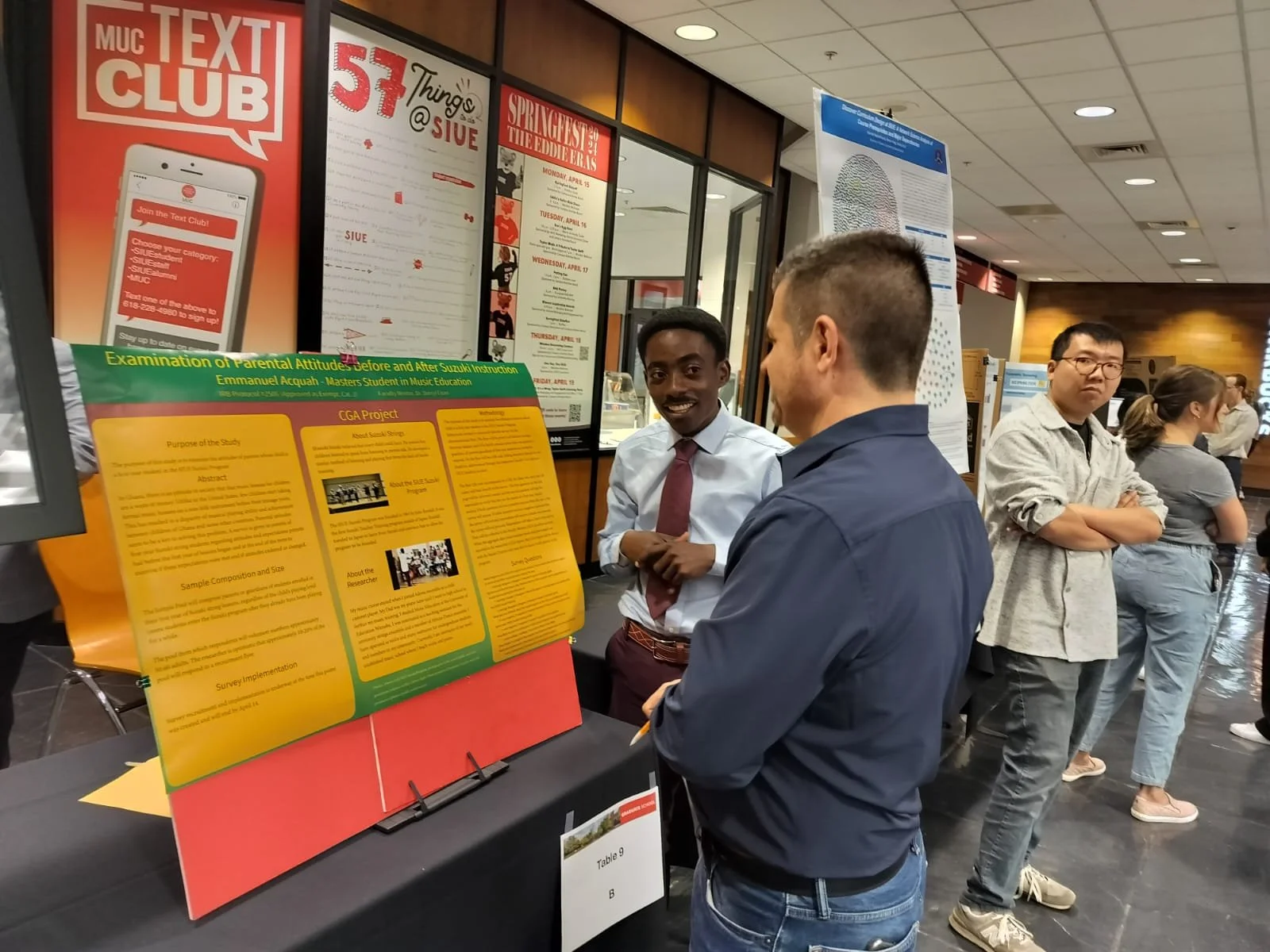 People attending a research poster presentation at an academic conference or event, with Prophakwa explaining the poster to another, posters, and conference signage visible in the background.